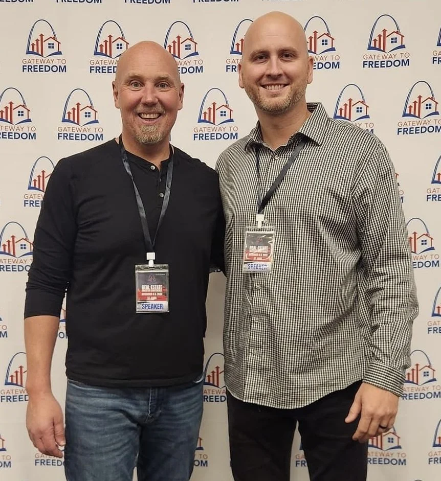 Two men standing side by side at a conference, smiling, wearing name badges, with a backdrop featuring a logo that includes a house, mountains, and the words "Gateway to Freedom."