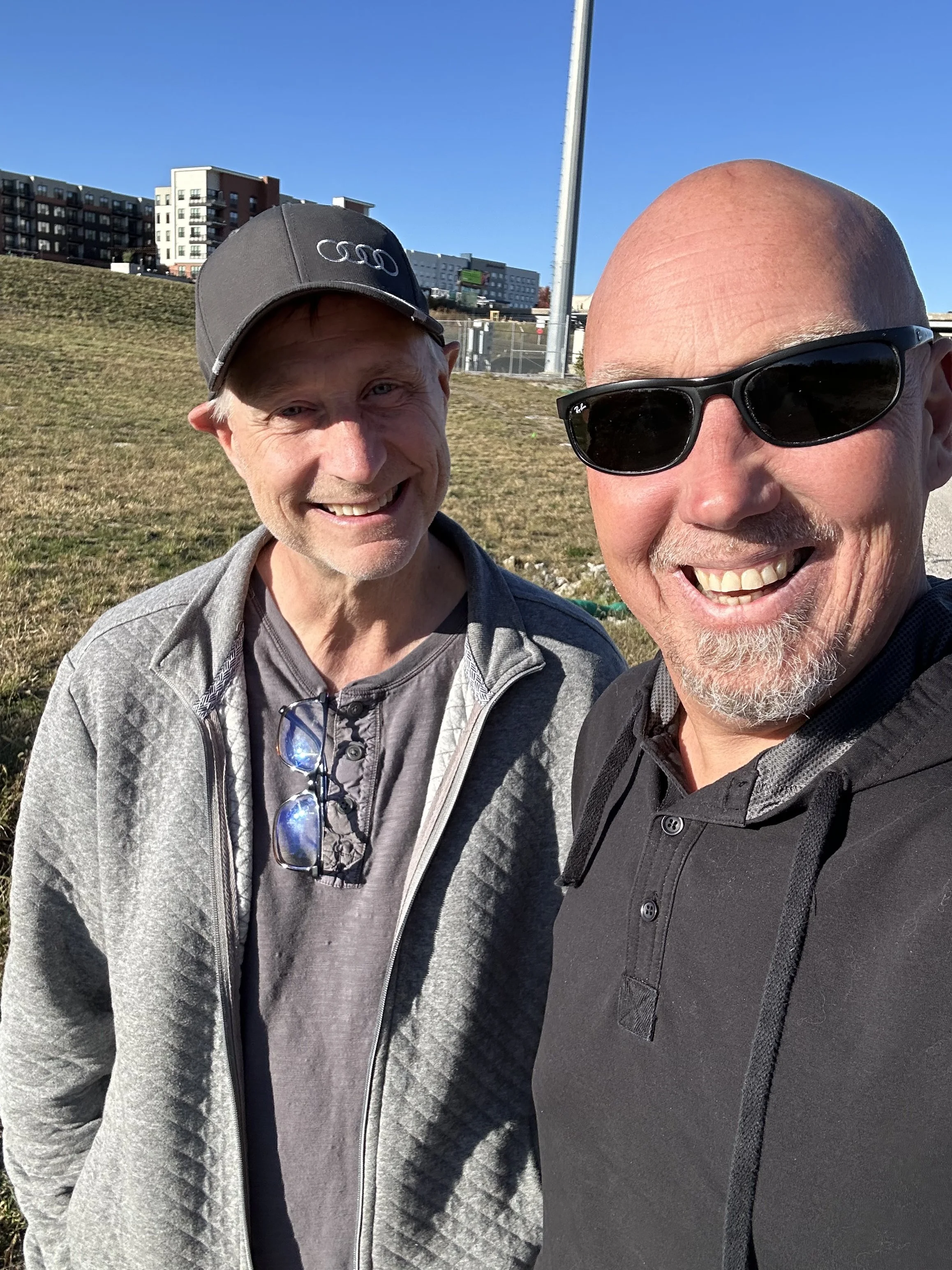 Two men smiling outdoors, one wearing a gray jacket and glasses hanging from his neck, the other wearing a gray cap with the Audi logo and black sunglasses, with an urban background and clear blue sky.