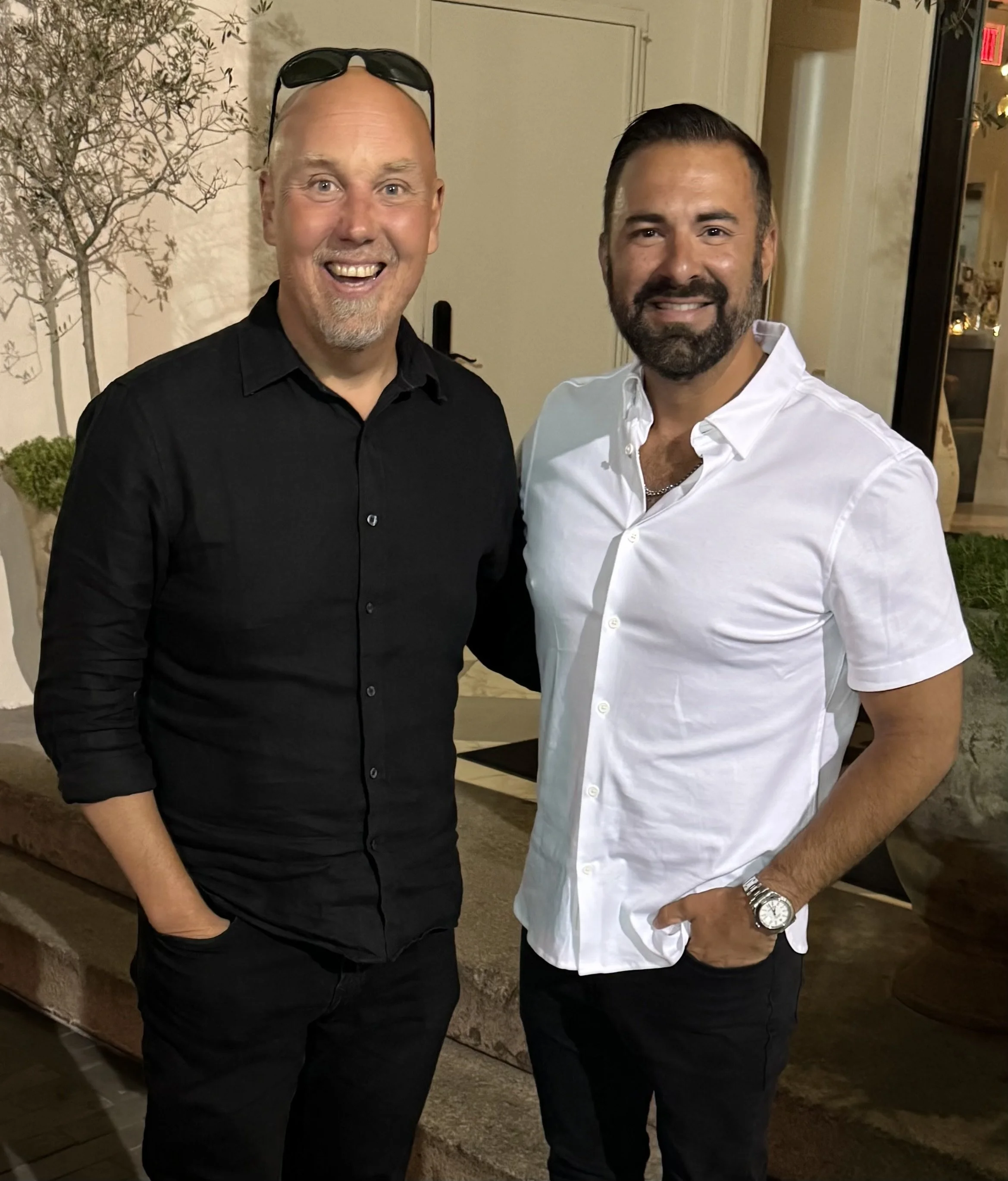 Two men smiling and standing close together outdoors at night, with a beige wall, plants, and a window behind them. The man on the left has a shaved head, sunglasses on his head, and is wearing a black shirt. The man on the right has dark hair, a beard, and is wearing a white shirt with rolled-up sleeves, a wristwatch, and a chain necklace.