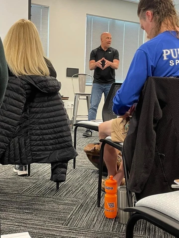 A man in athletic attire giving a presentation or speech to a group of people seated. The room has large windows with blinds, carpeted floor, and chairs. Some attendees have water bottles and backpacks.