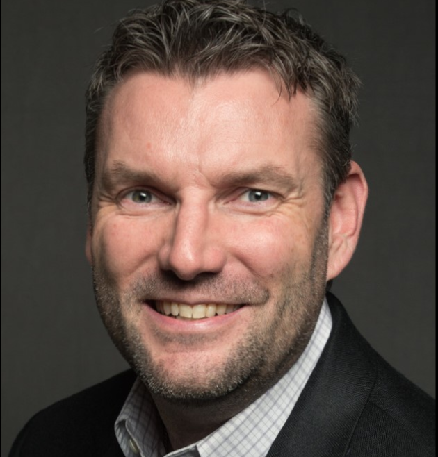 Close-up portrait of a smiling man with short, dark hair, a beard, blue eyes, wearing a black blazer and a light checkered shirt against a dark background.