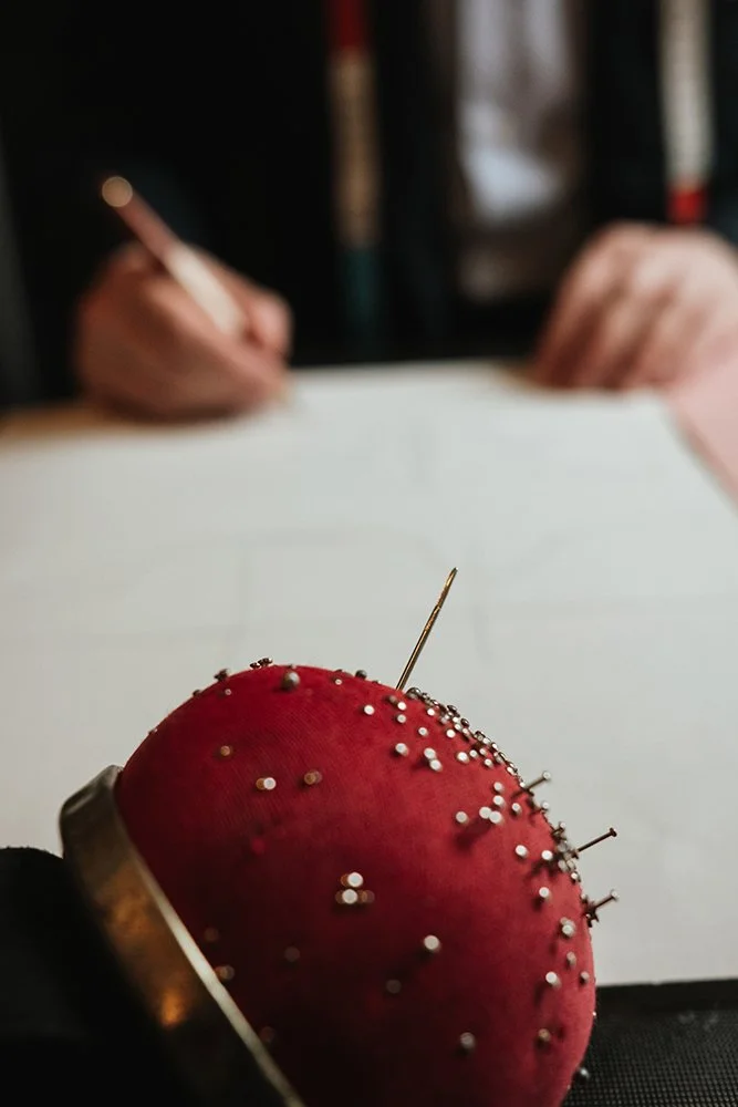 Une broderie en forme de fruit rouge avec des épingles, vue de près, avec une personne qui travaille sur un dessin ou un patron en arrière-plan.