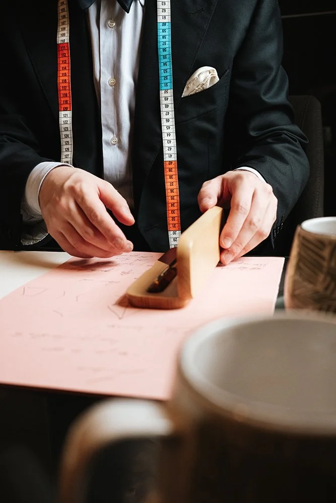 Une personne en costume noir et chemise blanche utilisant une règle de couture pour couper du papier rose, avec des tasses dans le premier plan.