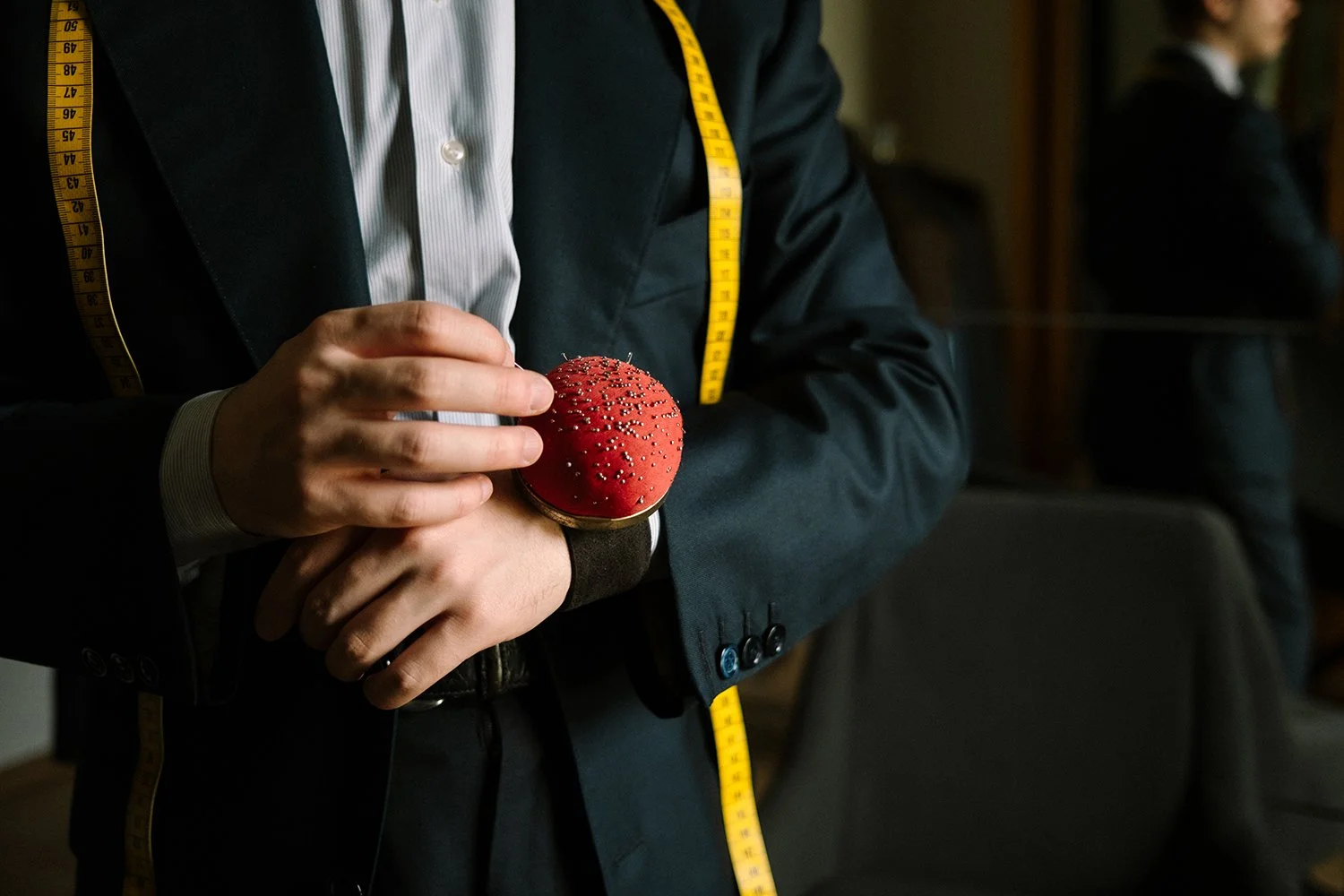 Une personne en costume noir avec un mètr enroulé autour du cou tient un porte épingle rouge.