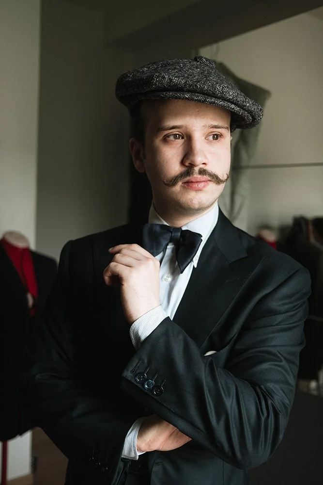 Homme avec une moustache, portant un costume noir, une chemise blanche, un nœud papillon et un béret gris, en train d'ajuster son nœud papillon.