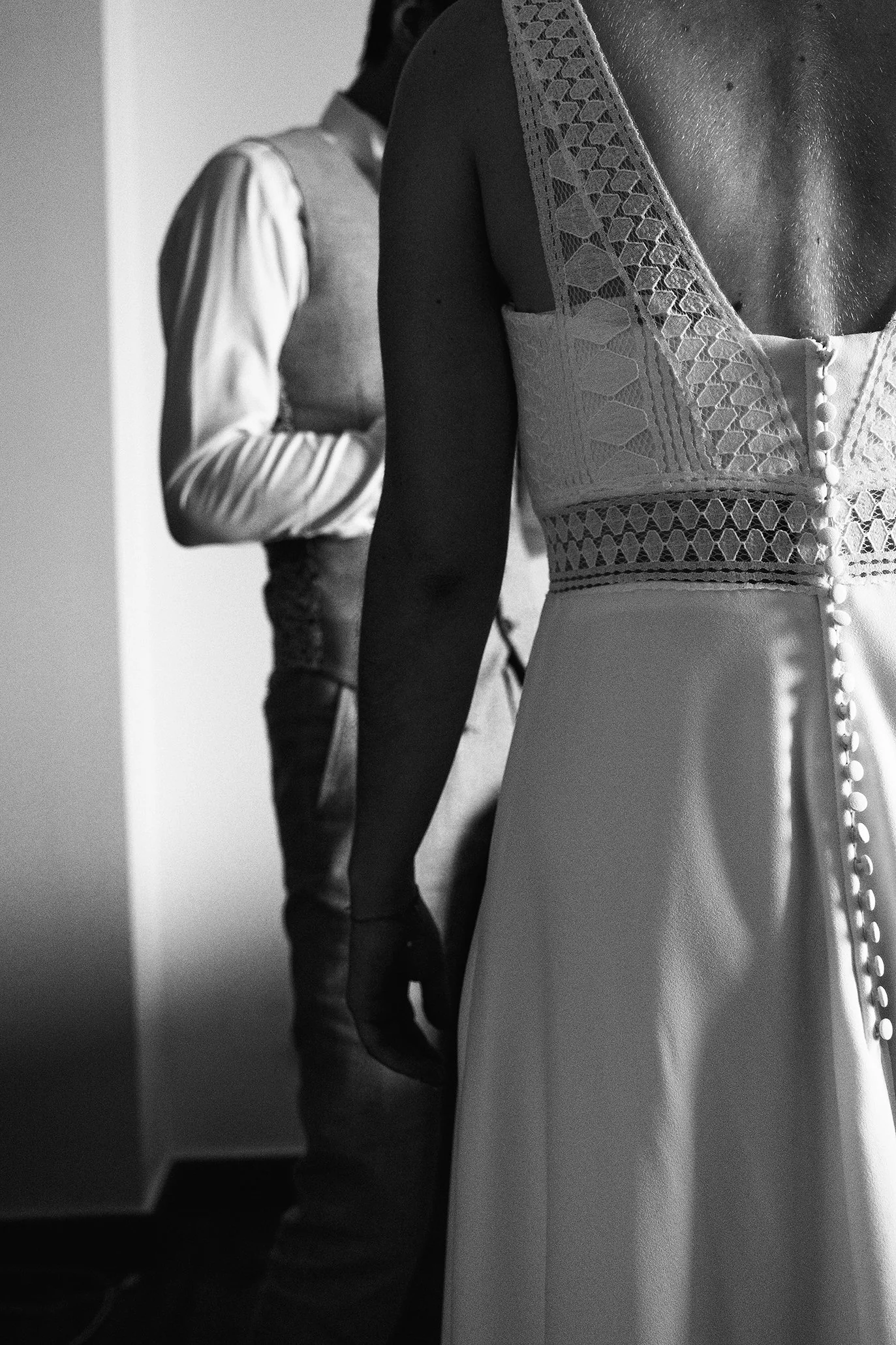 Photo en noir et blanc de deux personnes, une femme en robe avec détails en dentelle et boutons dans le dos, et un homme en veste, dans un espace intérieur.