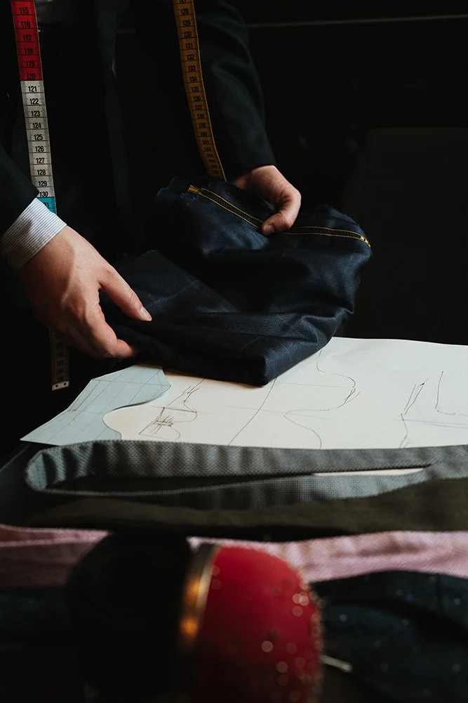 Une personne en costume utilisant un mètre-ruban pour mesurer un vêtement ou un patron sur un bureau. Présence également d'une paire de ciseaux, d'une sacoche en toile et d'un crayon ou filets pour dessin.