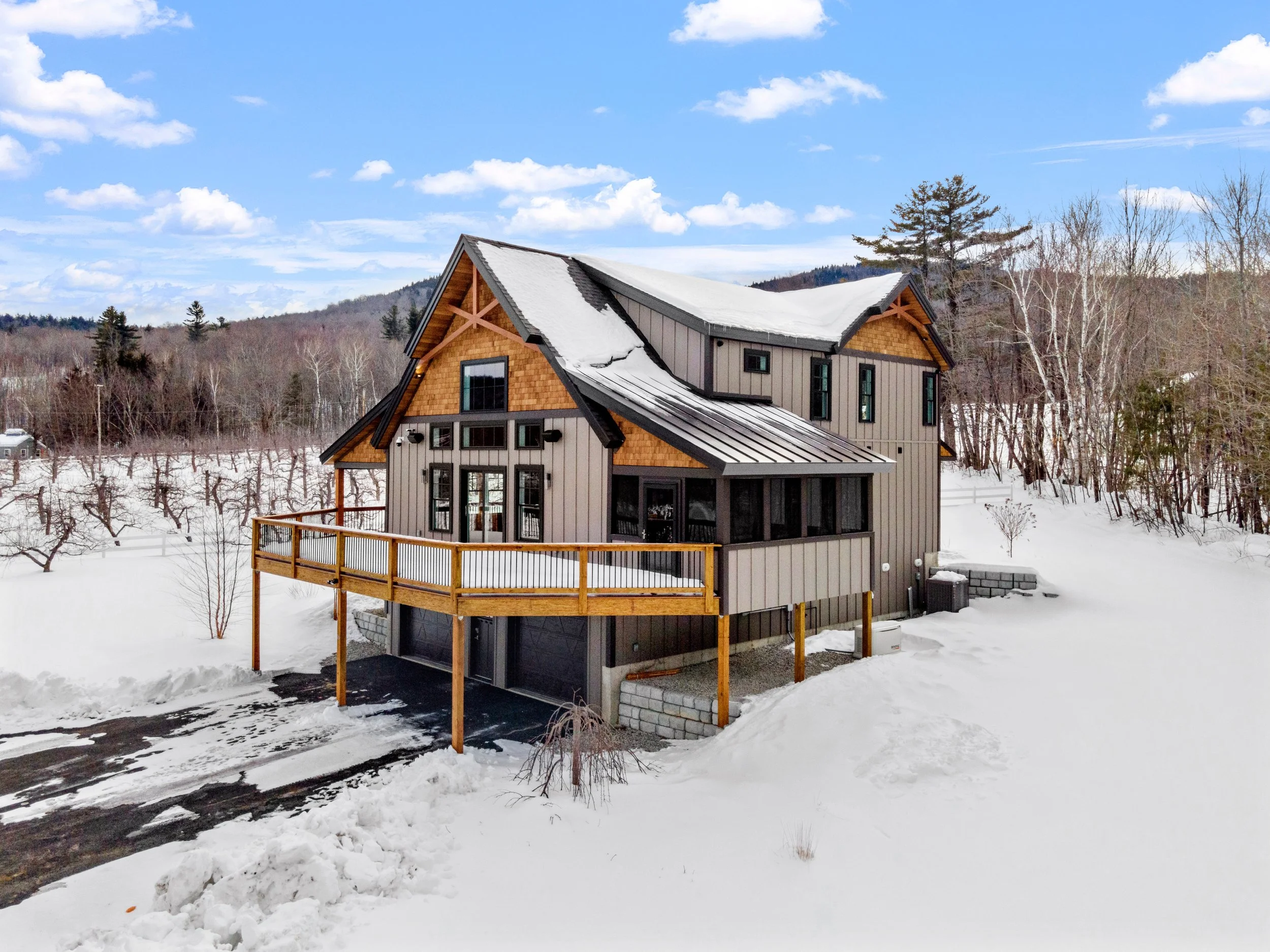 Modern cabin-style house with snow-covered roof and raised wooden deck, surrounded by a snowy landscape and trees, under a blue sky with clouds.