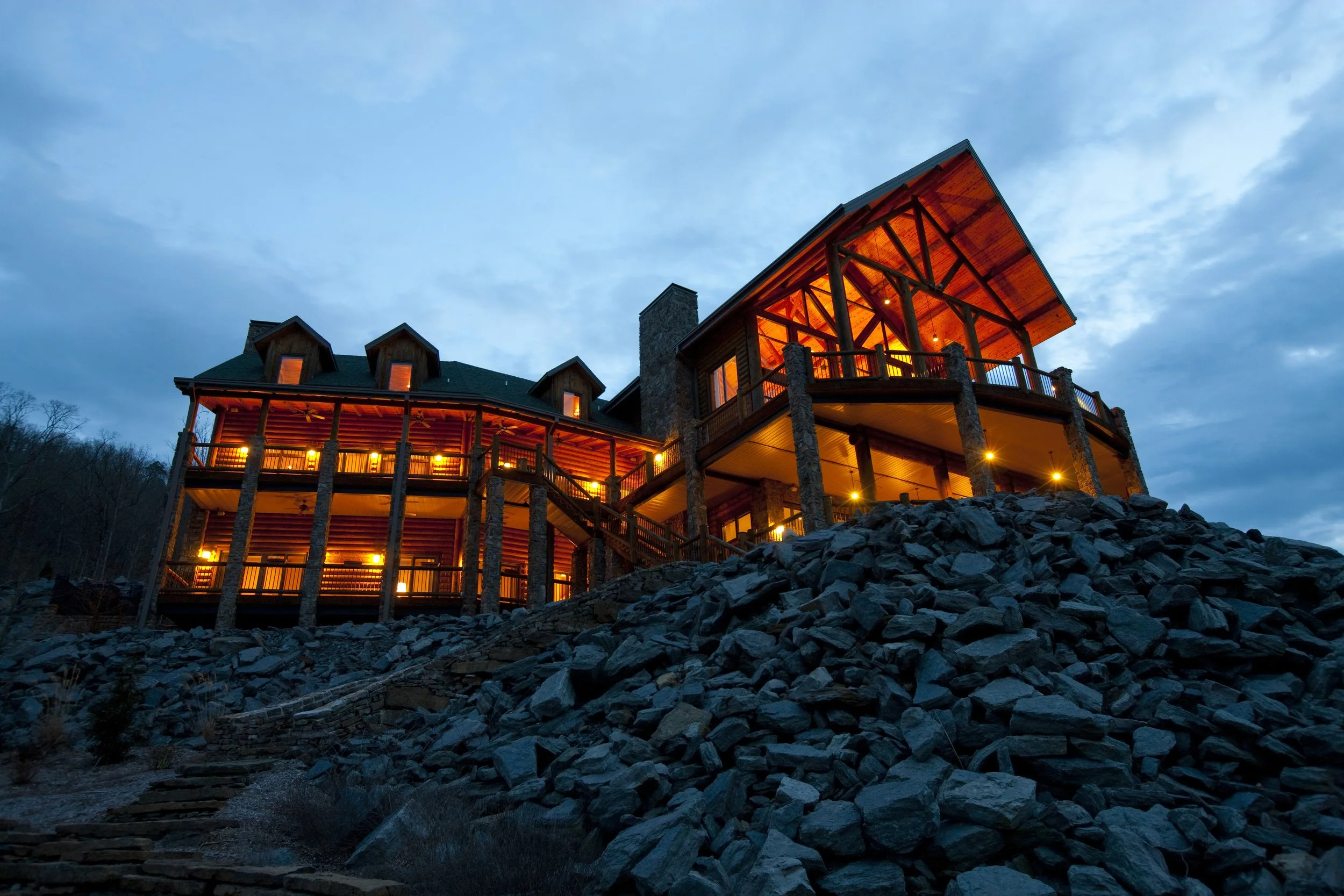 Large log cabin with stone pillars and warm lighting at dusk, surrounded by rocky terrain.