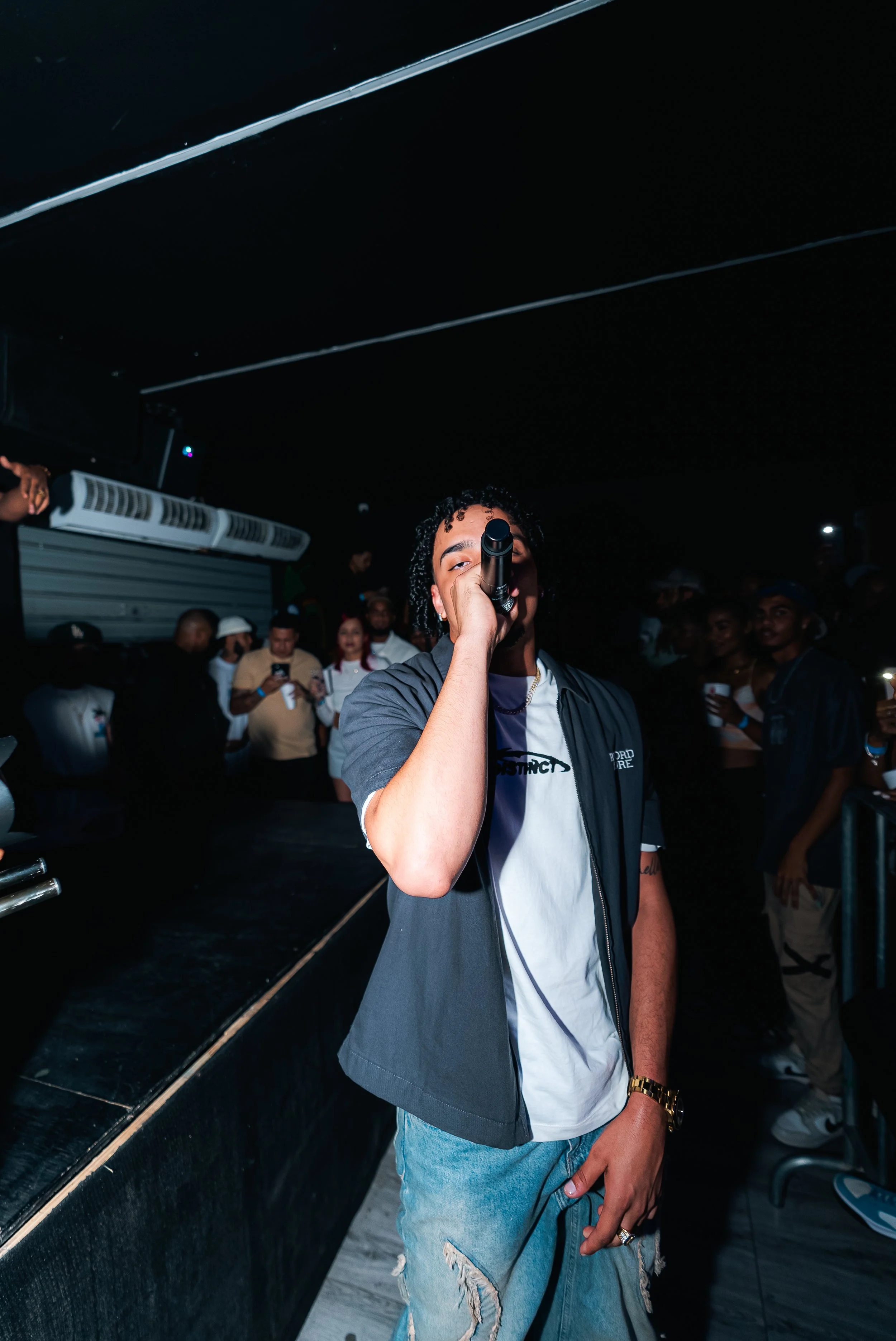A man holding a microphone performing on stage with an audience watching in a dimly lit venue. He is wearing a casual outfit including distressed jeans, a white t-shirt, and a dark open shirt. People in the background are taking photos and videos wit