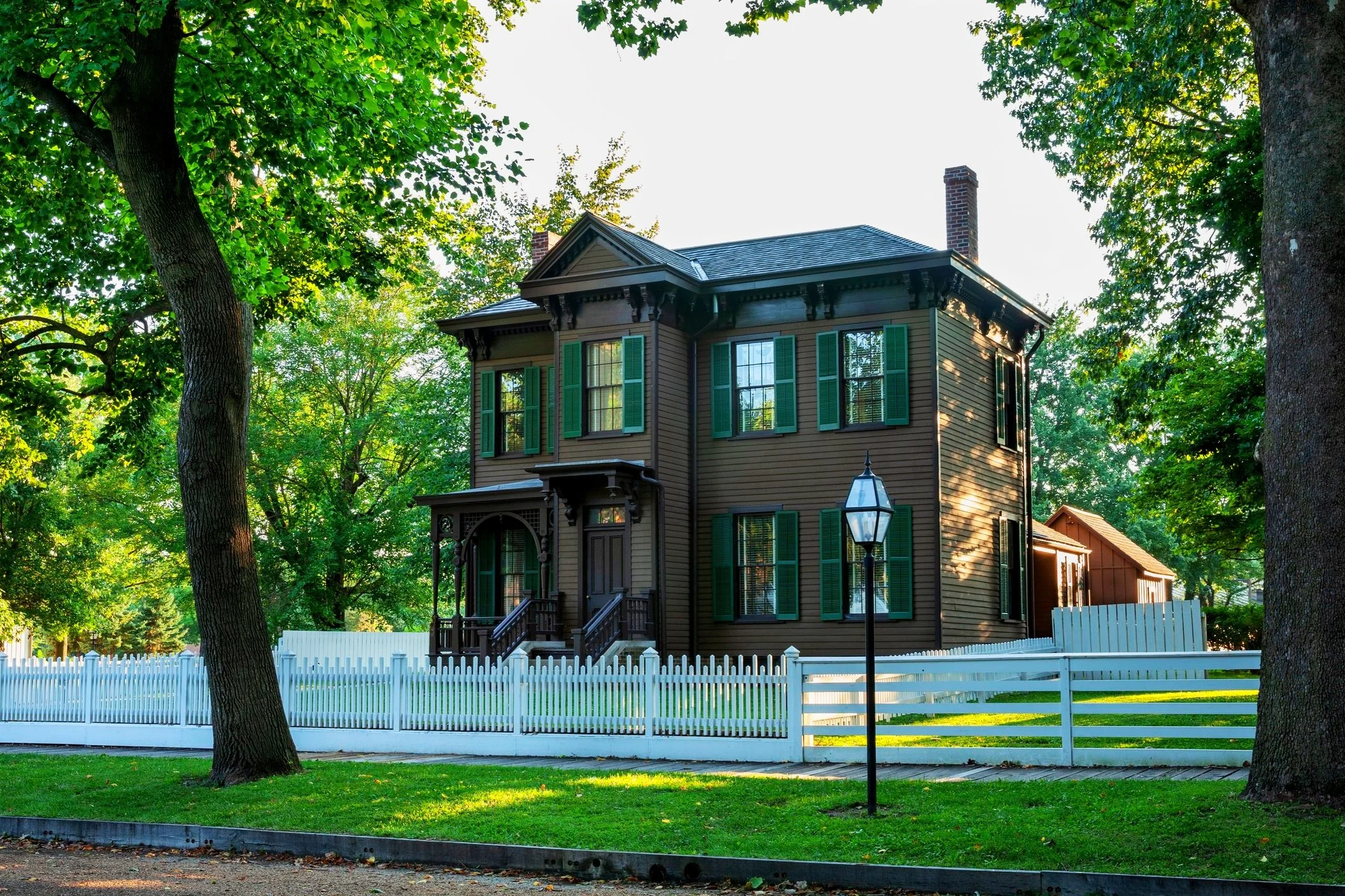 Historic two-story brown house with green shutters, surrounded by trees and a white picket fence, featuring a black lamppost on a grassy lawn.