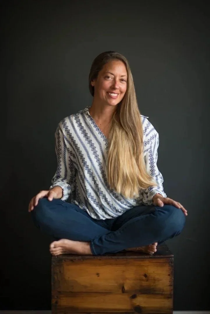 A woman with long blonde hair sitting cross-legged on a wooden box against a dark gray background, smiling at the camera.
