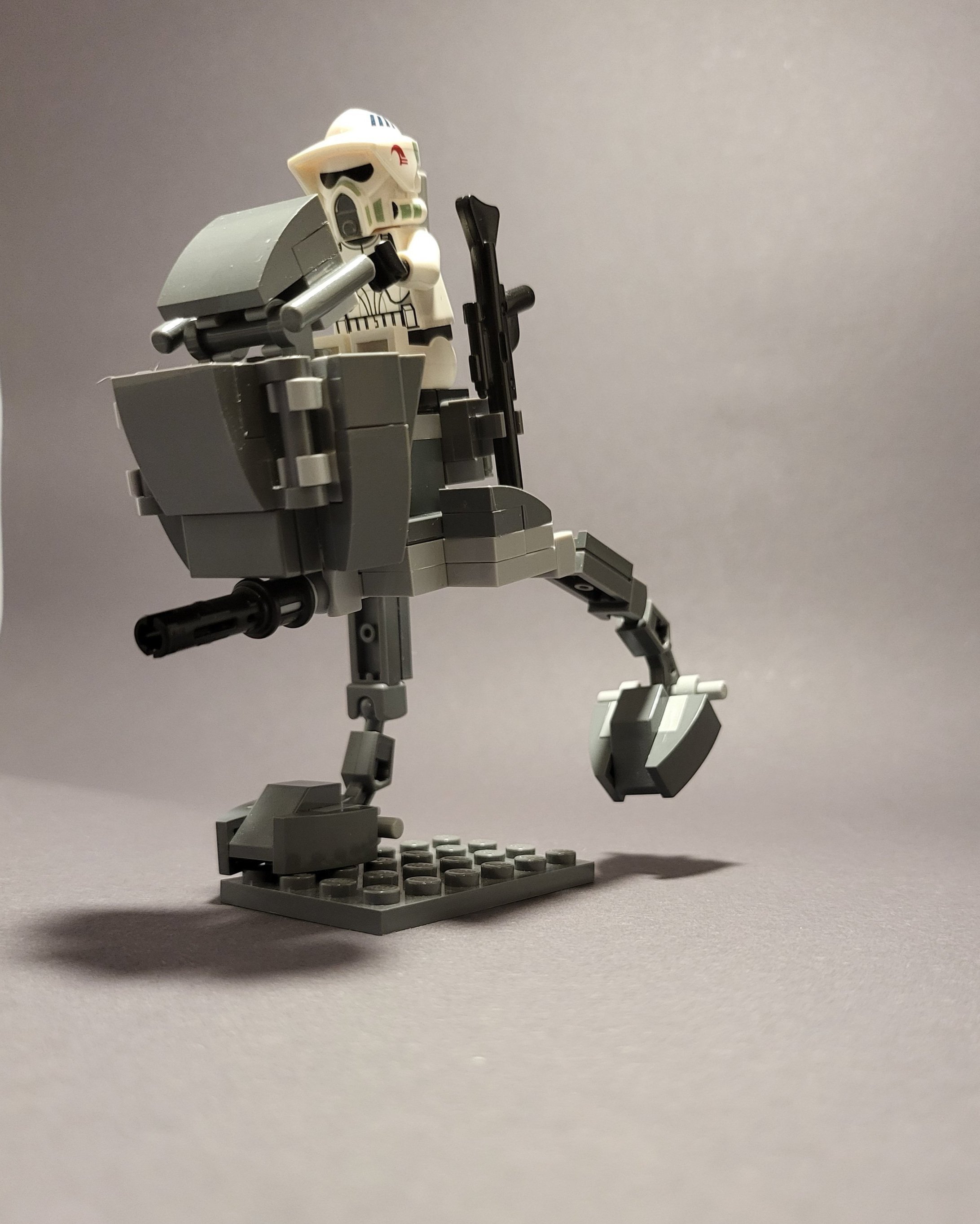 LEGO figure with helmet riding a gray mechanical walker on a gray background.