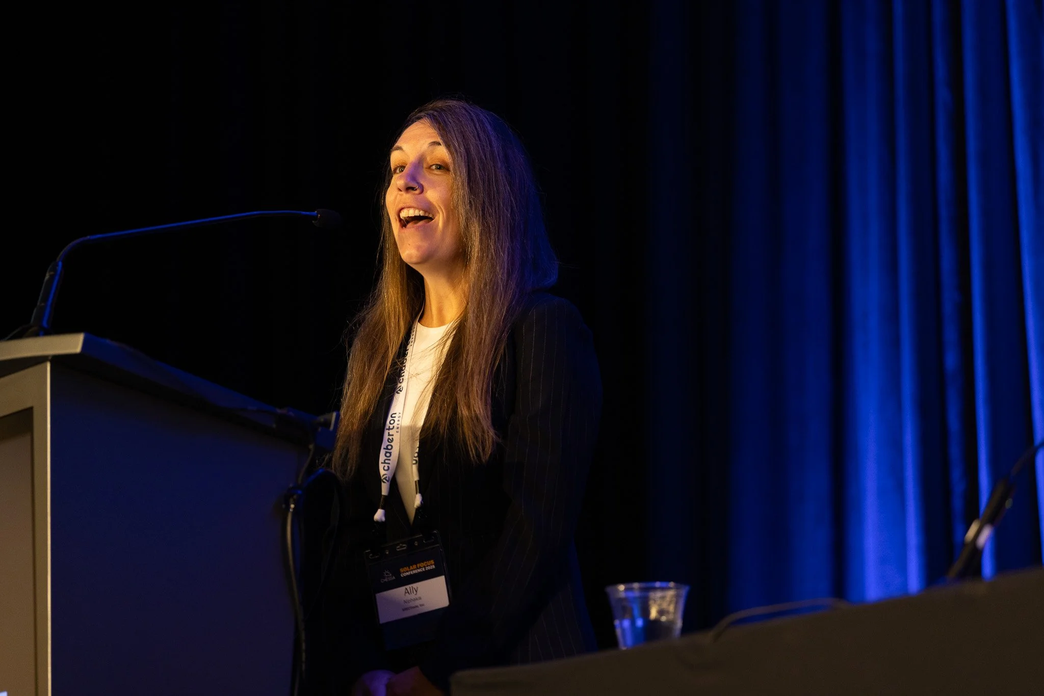 A woman giving a speech at a podium on stage, with colorful stage lighting and blue curtains in the background