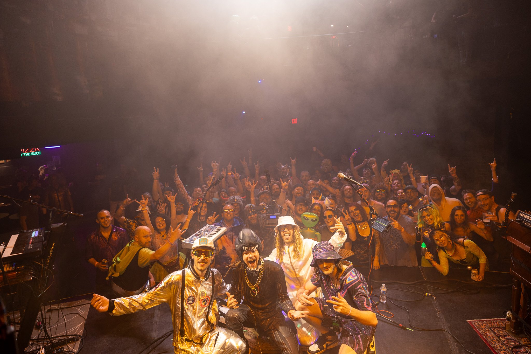 Group of performers on stage in colorful costumes with musical instruments, smiling and posing for the camera, while a large audience behind them makes peace signs and cheers in a lively concert setting.