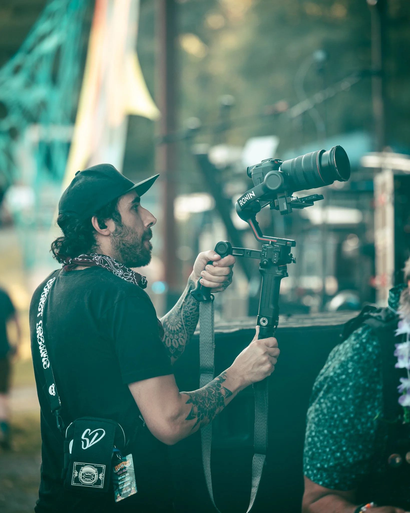 A man operating a camera stabilizer with a large lens, wearing a black cap, black t-shirt, and a black scarf with white pattern, outdoors during daytime.