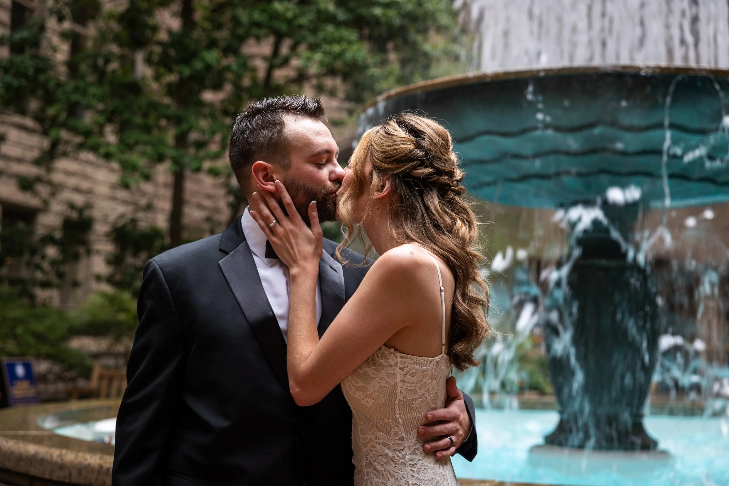 A newlywed couple kissing in front of a fountain amidst a lush outdoor setting.
