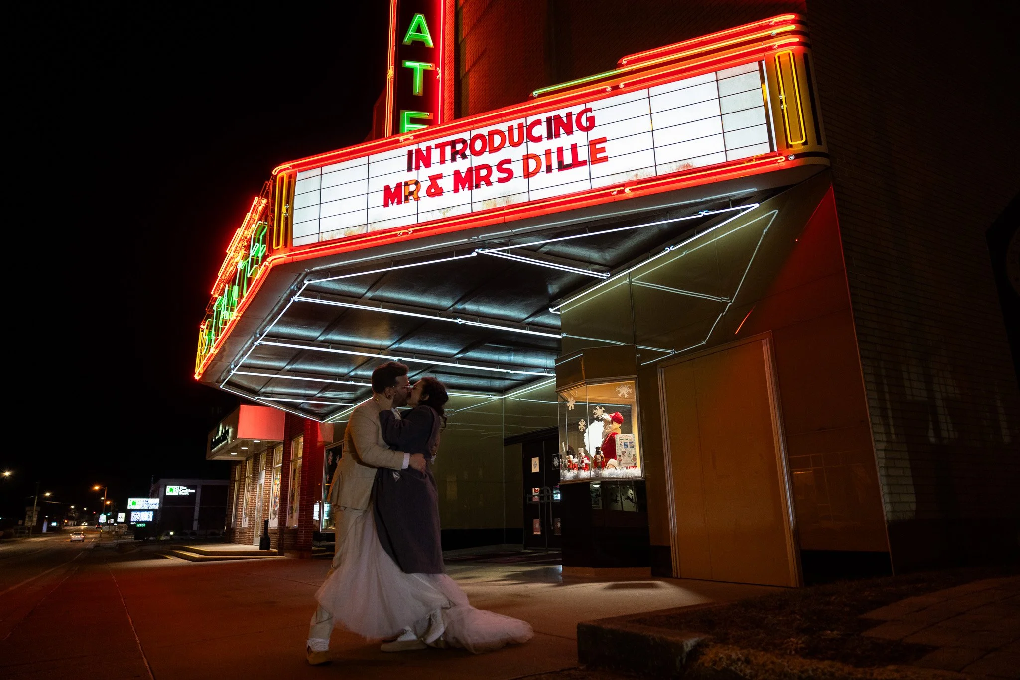 A couple dressed in wedding attire sharing a kiss in front of a brightly lit theater marquee at night, with festive holiday decorations inside the theater window.