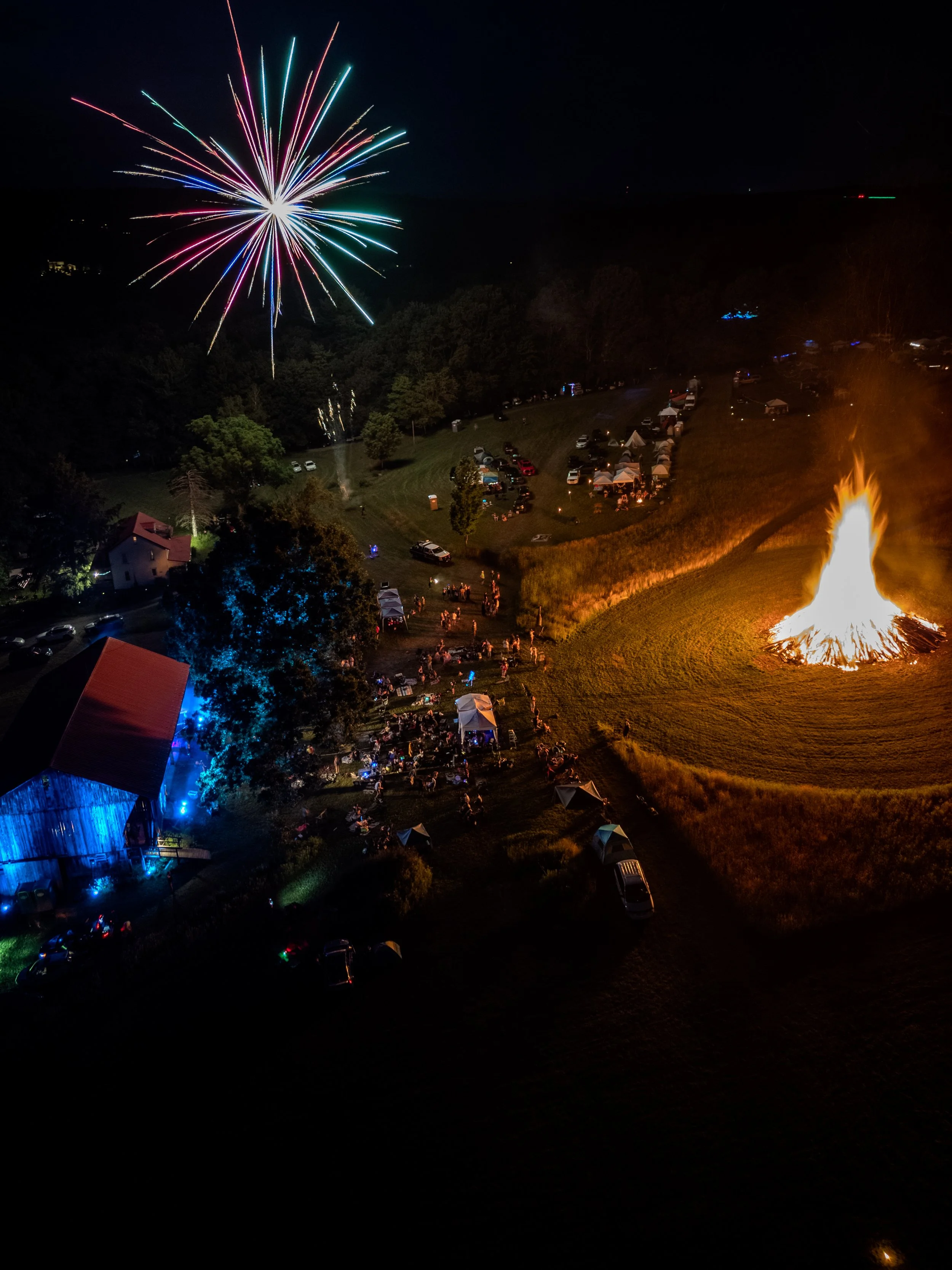 Fireworks exploding in the night sky over a large outdoor gathering with tents, cars, and a large bonfire.
