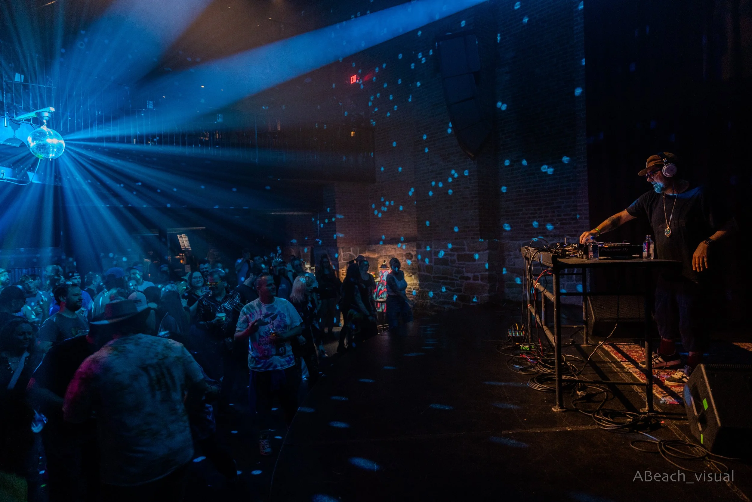 A DJ performing on stage with blue disco lights illuminating a crowd of people dancing at a nightclub or concert venue with brick walls.
