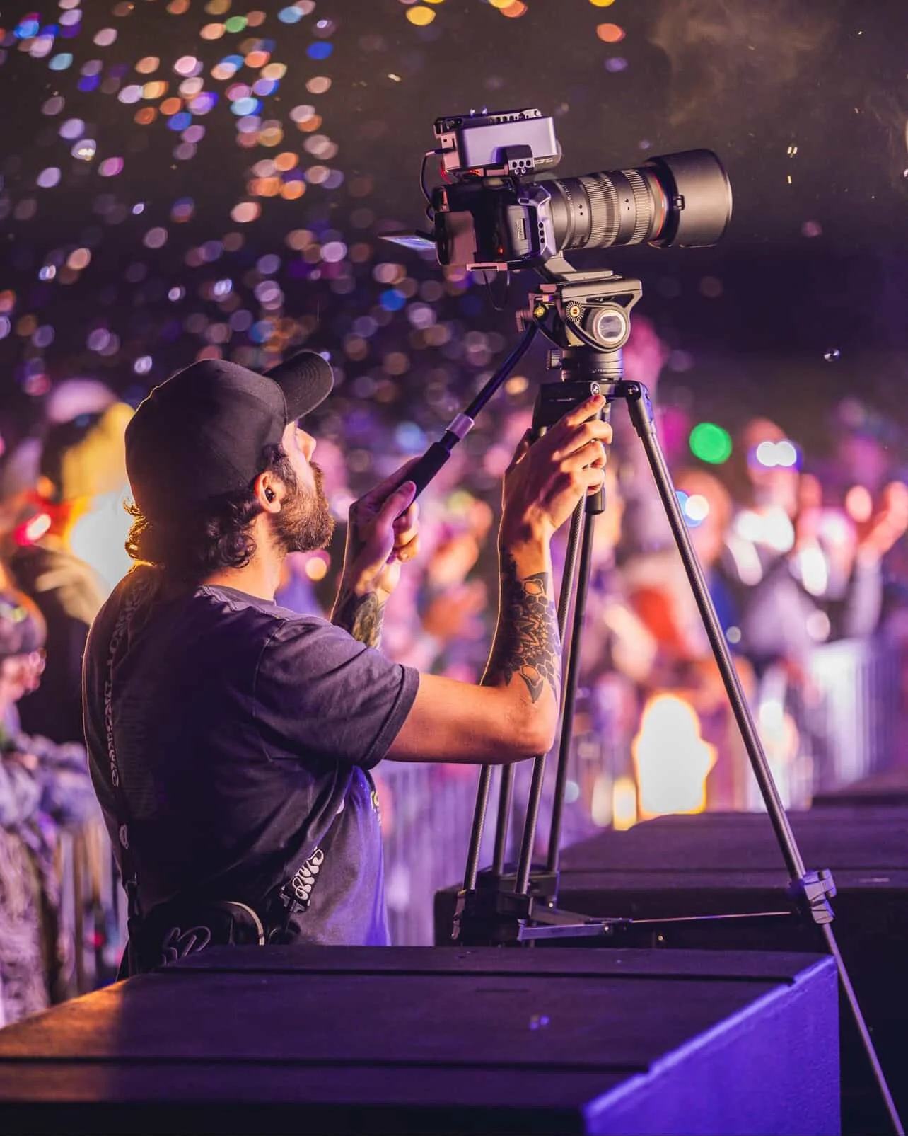 A man with a beard, wearing a black cap and black t-shirt, operating a professional video camera on a tripod at an outdoor event during nighttime. The background is filled with colorful, out-of-focus lights and a crowd of people.