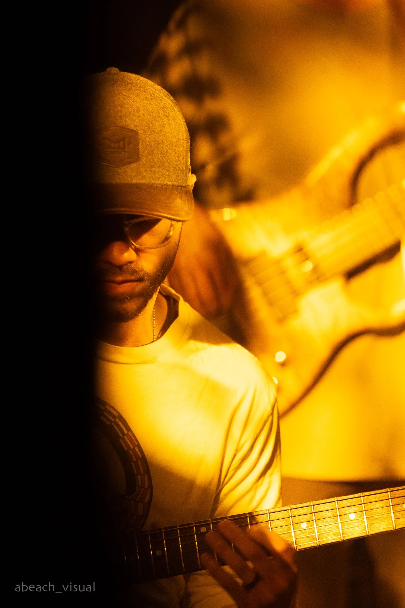 A young man wearing glasses, a cap, and a beige shirt, playing an acoustic guitar in a dimly lit environment with yellow-orange lighting.