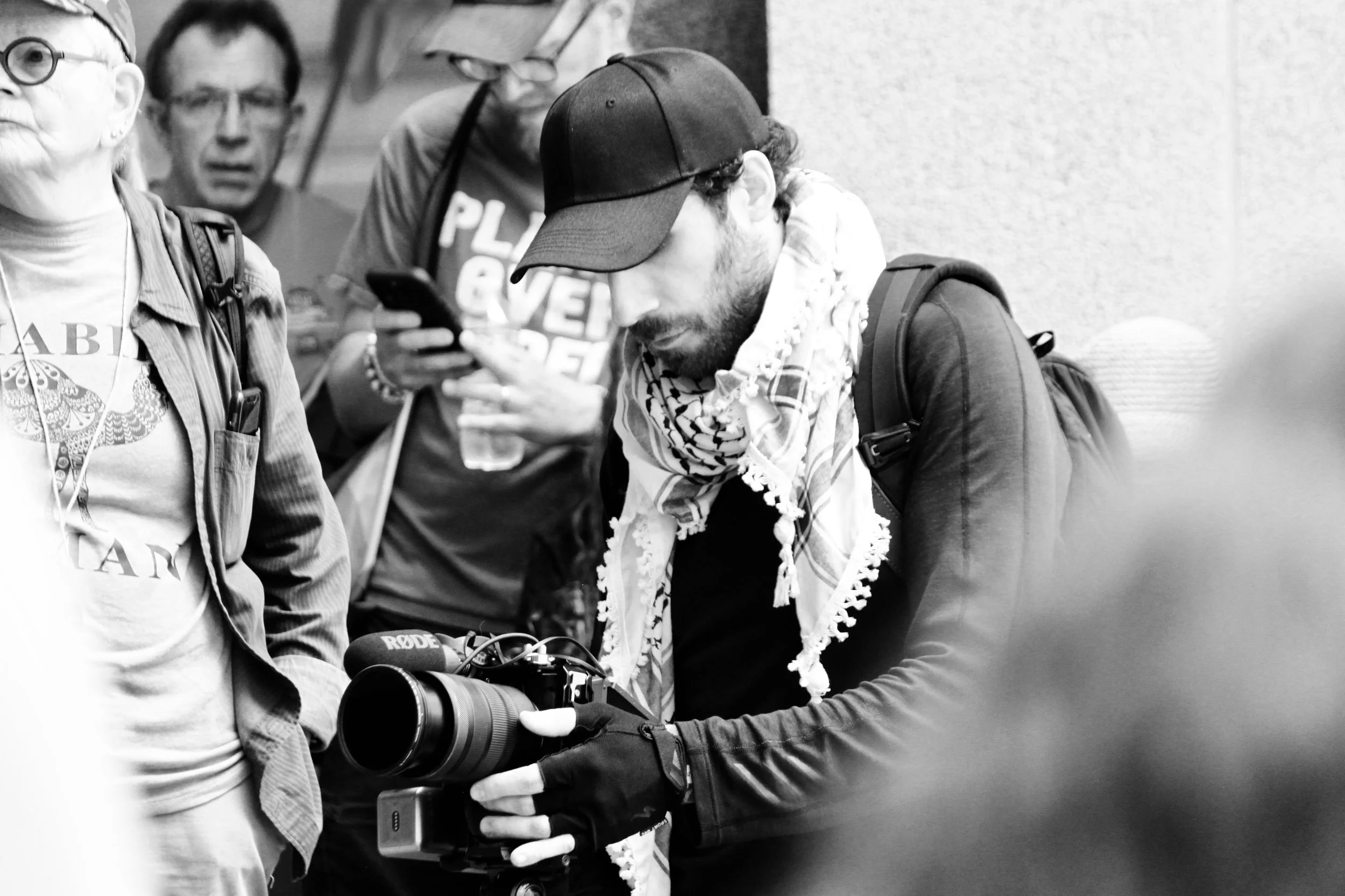 Group of people, including a man with a cap, beard, and scarf, holding a camera and looking at it, while others around him focus on their phones or observe.