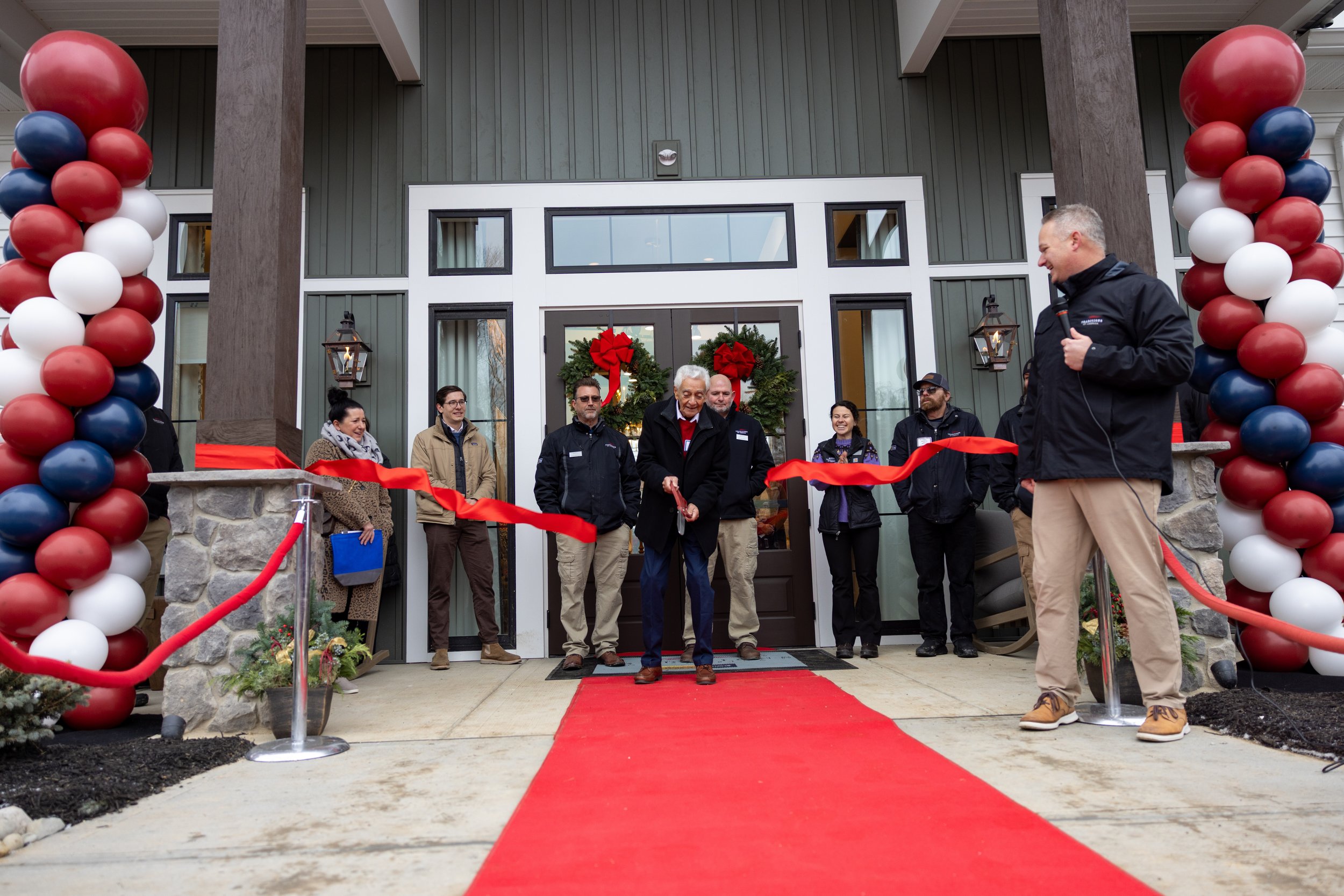 A ribbon-cutting ceremony taking place in front of a house decorated with Christmas wreaths and balloons, with several people present and a man speaking into a microphone.