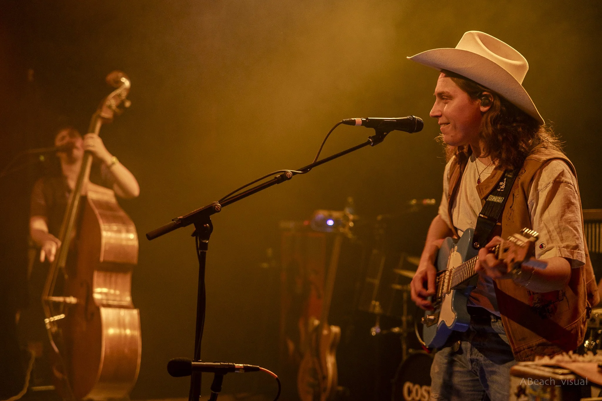 Musicians performing live on stage, one with an acoustic guitar wearing a cowboy hat, and another playing a double bass in the background.