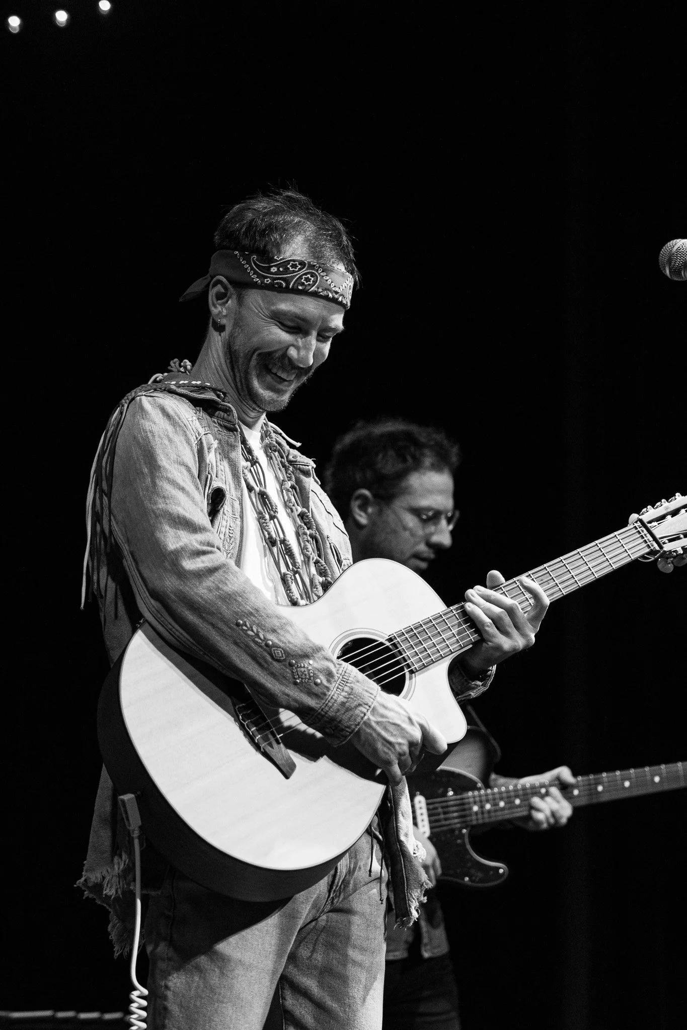 Two musicians playing guitars on stage, one in the foreground smiling and wearing a bandana, the other slightly behind in the background.
