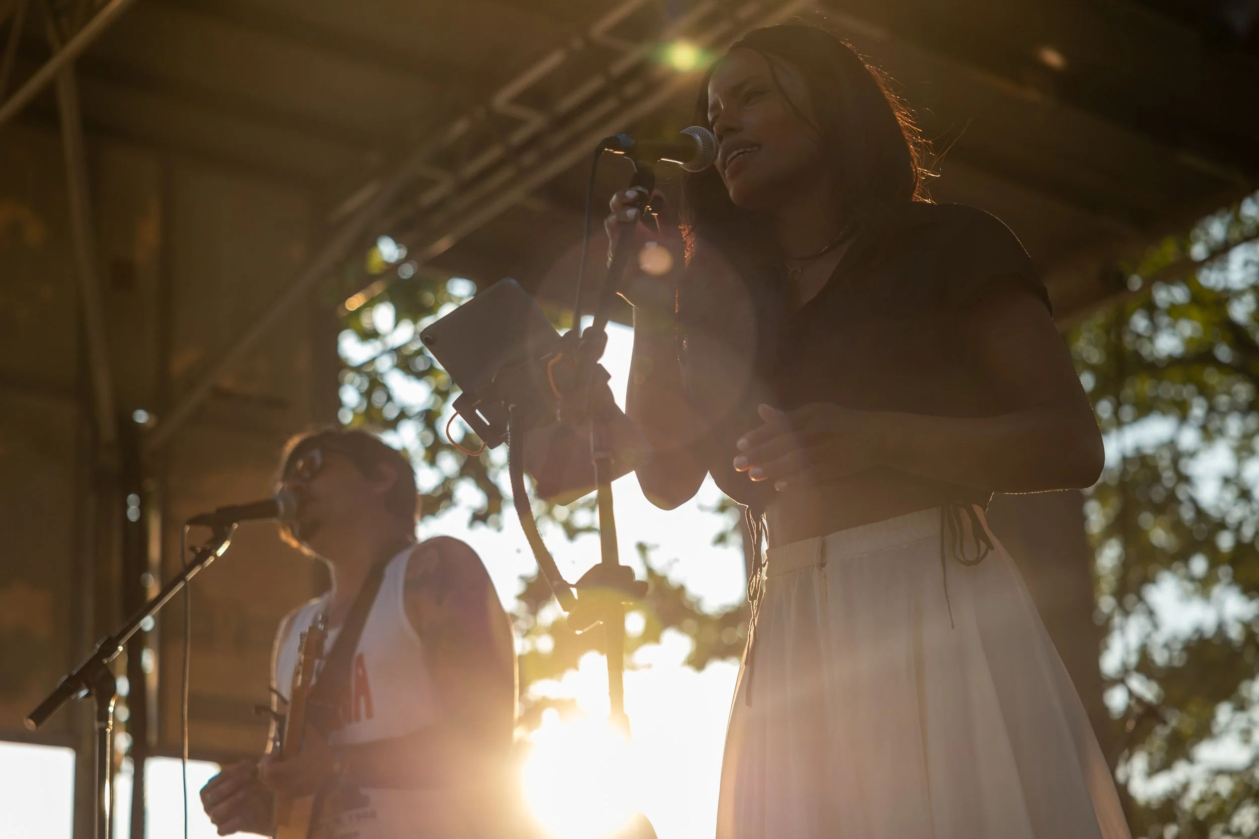Two women performing on stage outdoors during sunset, with one woman singing into a microphone and the other playing an instrument.