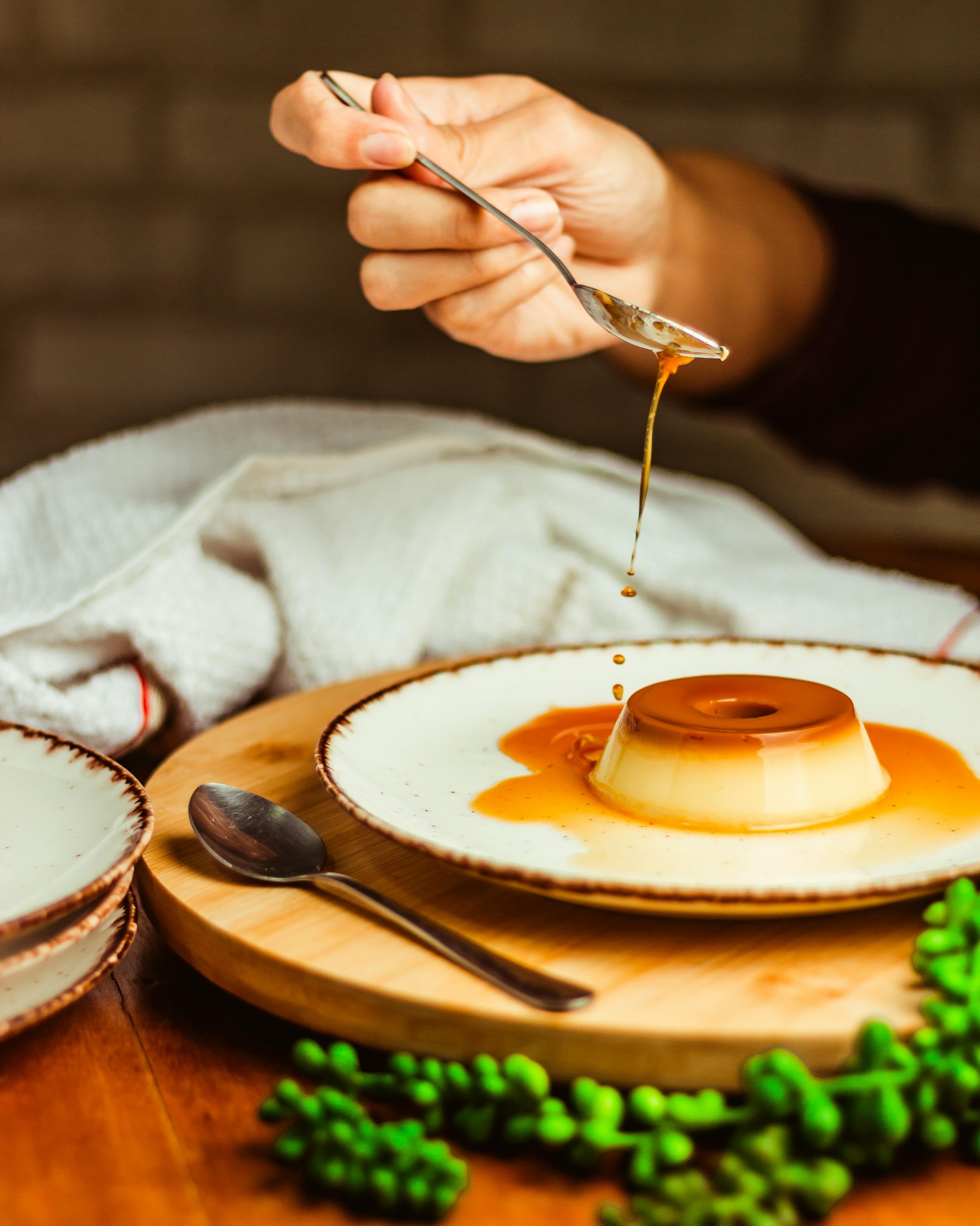 Close-up of a person holding a spoon with caramel sauce, pouring it into a flan on a white plate, side view showing caramel drizzling.