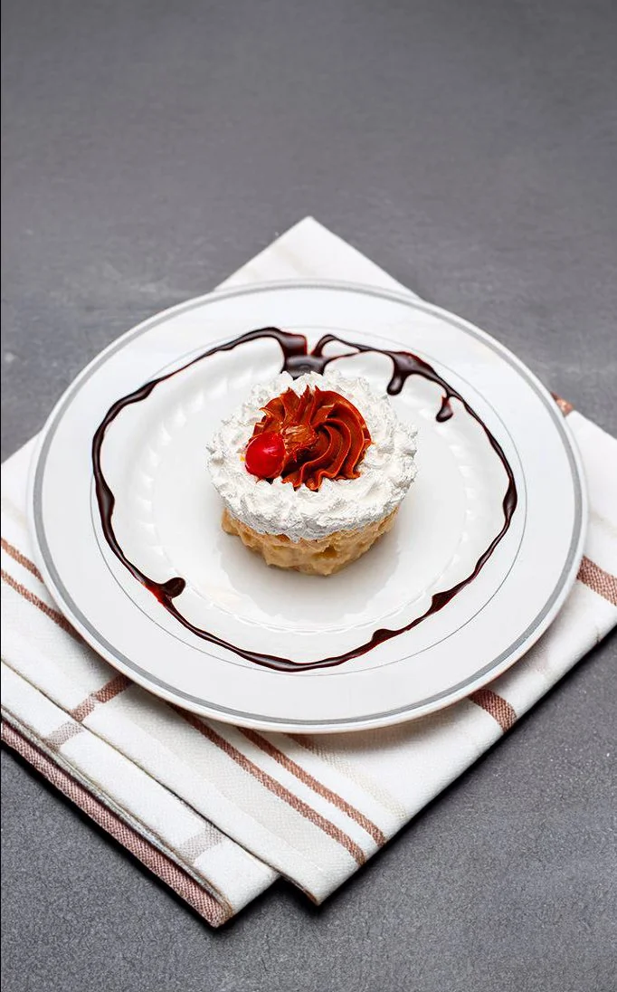 A beautifully presented cuatro leches cake topped with whipped cream, a dollop of caramel, and a bright red cherry, served on a white plate with a chocolate drizzle. The dessert sits on a folded cloth napkin, adding an elegant touch to the setting