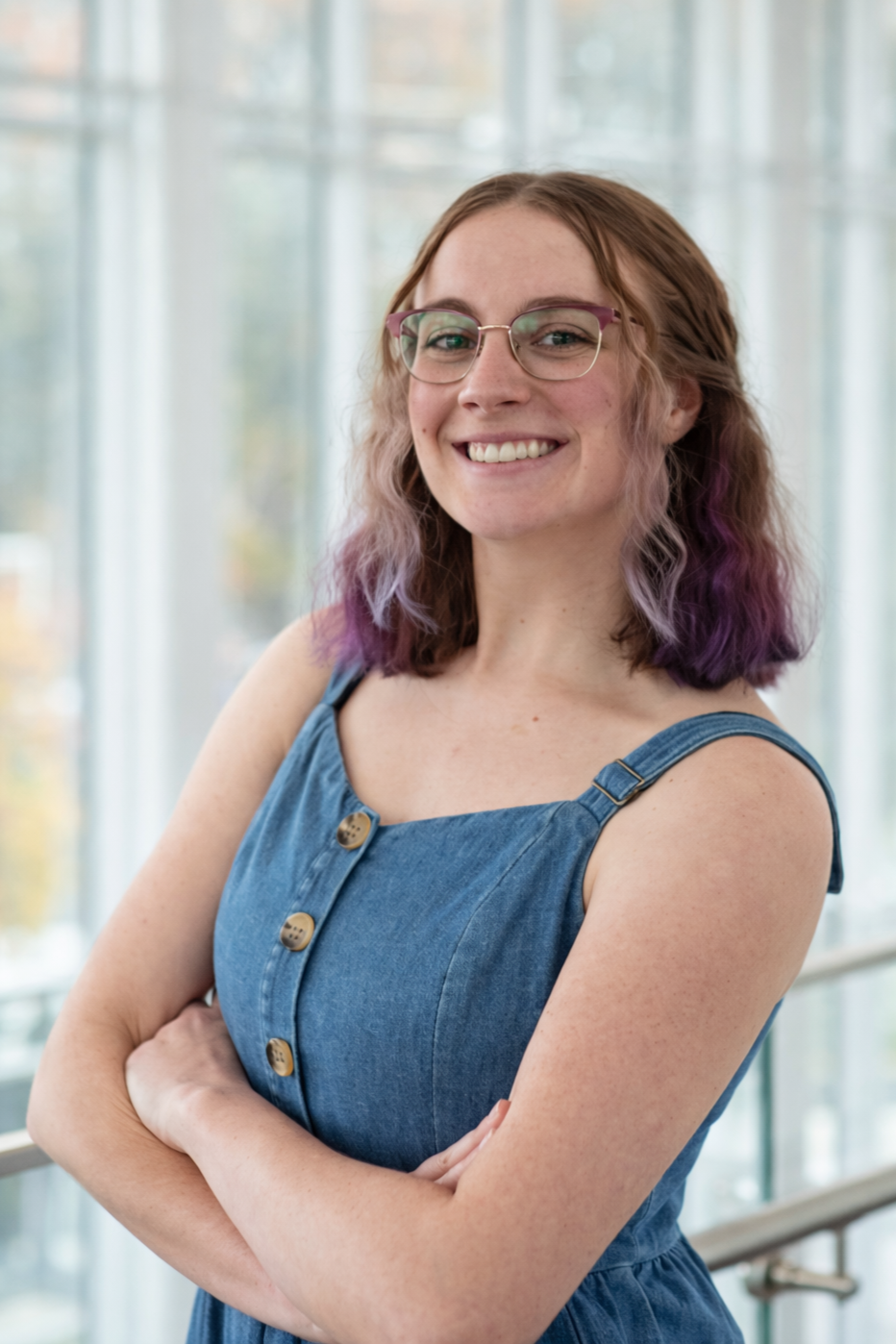 Young woman with purple curly hair, smiling, wearing a black graduation gown and earrings.
