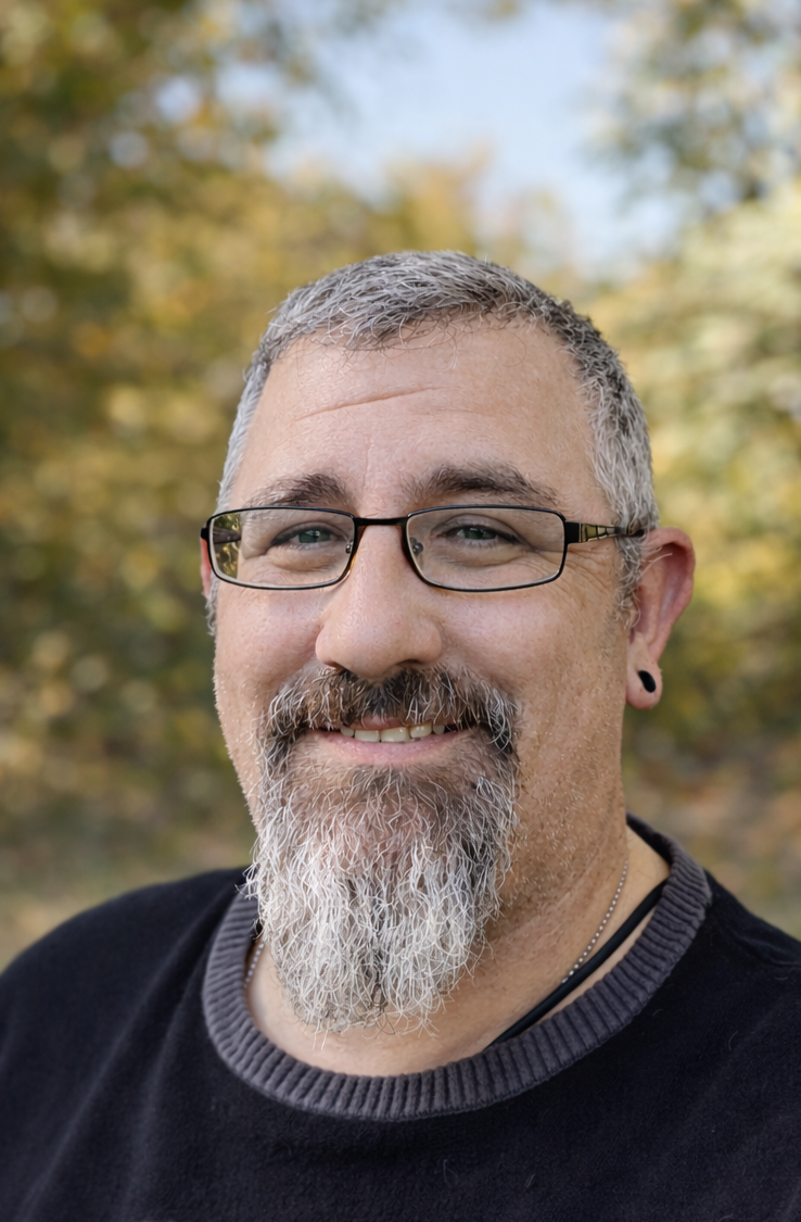 A man with glasses, a beard, and short hair, wearing a blue and white plaid shirt, standing outdoors against a brick wall.