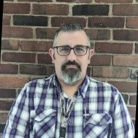 A man with glasses, a beard, and short hair, wearing a blue and white plaid shirt, standing outdoors against a brick wall.