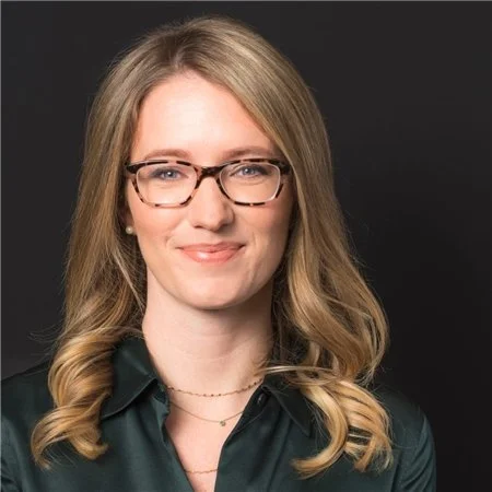 Portrait of a woman with blonde hair, glasses, and a dark green blouse, smiling against a black background.