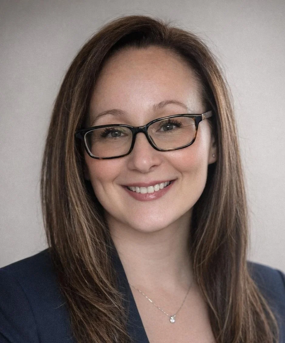 A woman with shoulder-length brown hair, wearing black-rimmed glasses, smiling, dressed in a dark blazer, and wearing a delicate necklace with a small pendant.