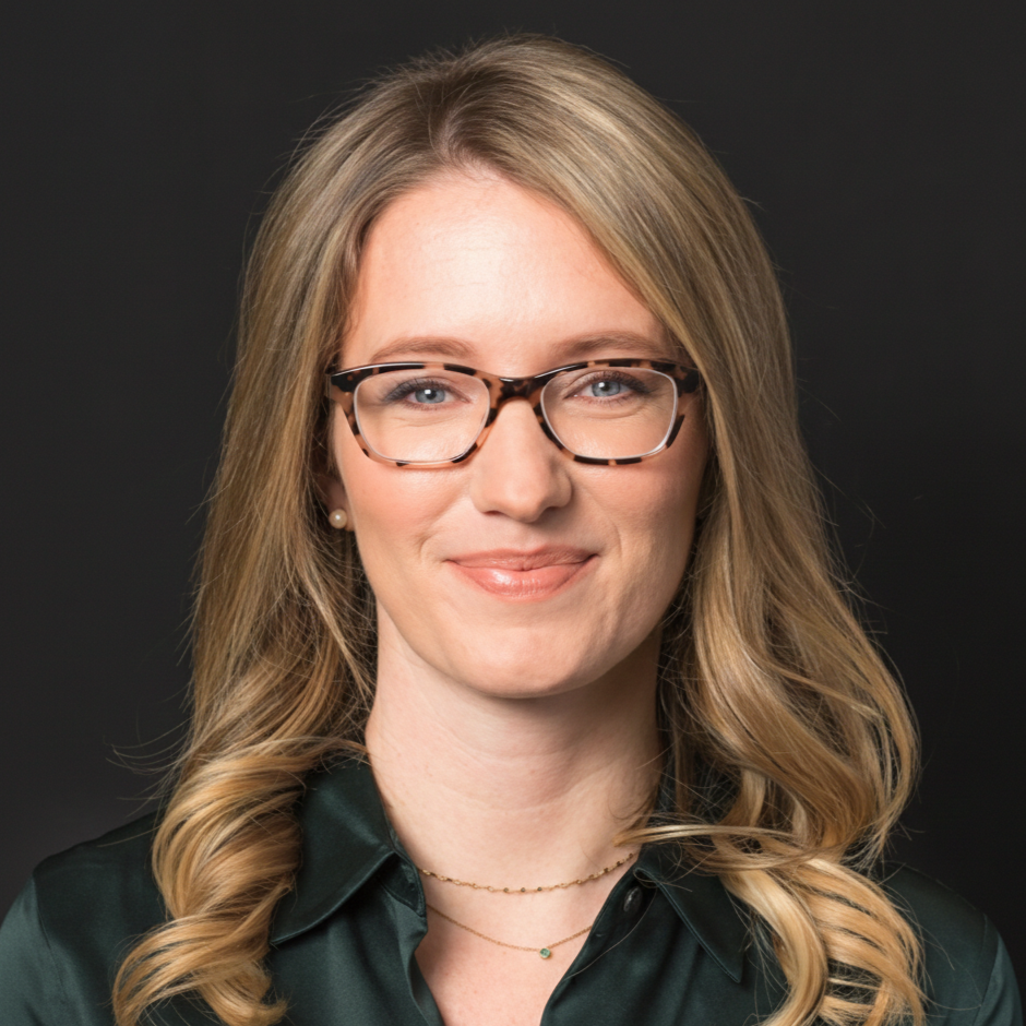 Portrait of a woman with blonde hair, glasses, and a dark green blouse, smiling against a black background.