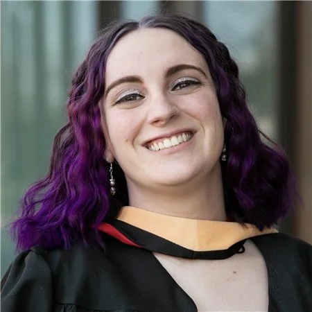 Young woman with purple curly hair, smiling, wearing a black graduation gown and earrings.