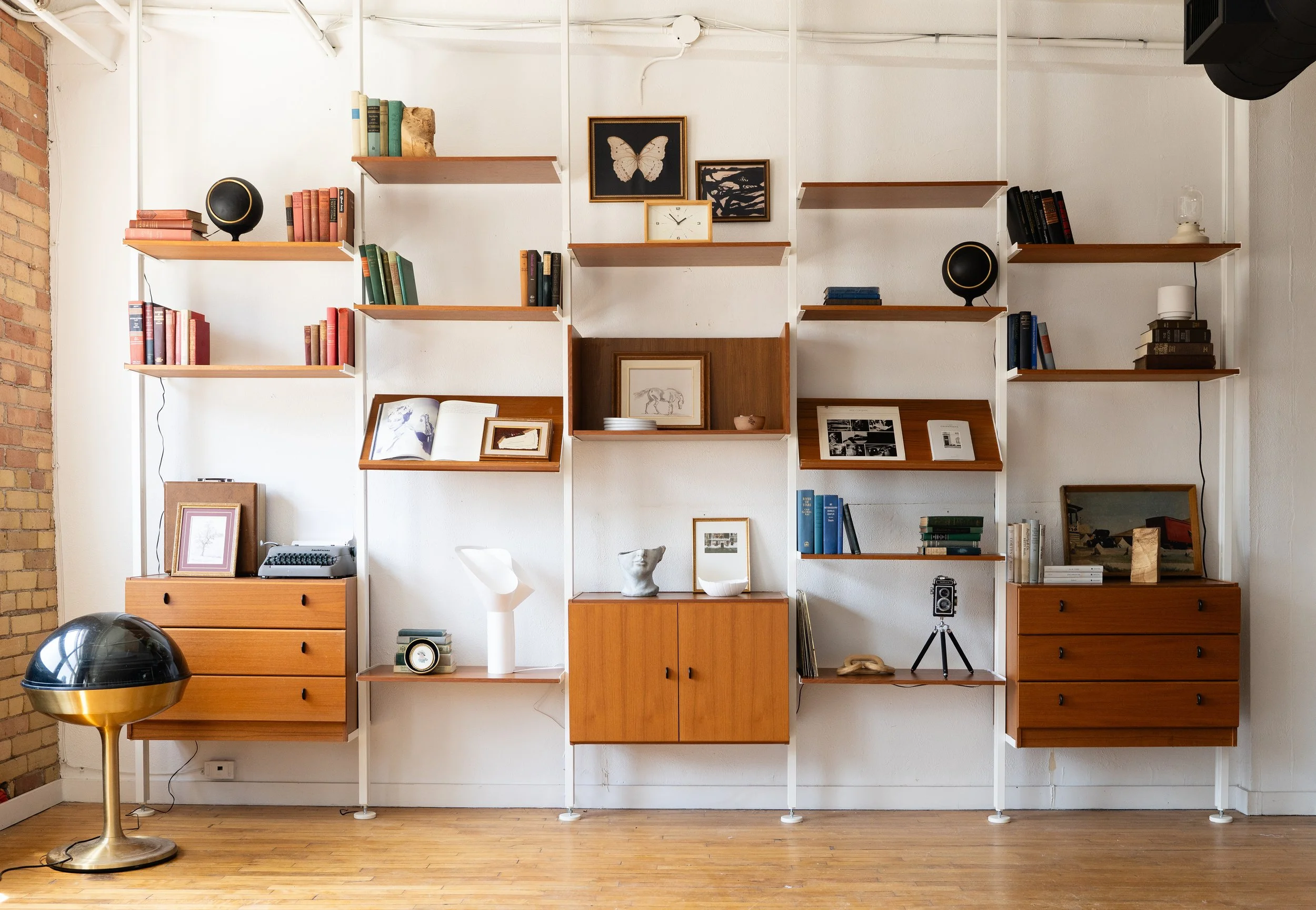 A modern interior with wood and white metal shelving on a white wall displaying books, framed art, and decorative objects. A vintage typewriter, a white bust, and framed photographs sit on the lower shelves. A black and gold globe and a black speaker are also part of the decor, alongside a golden lamp and hardwood floor.