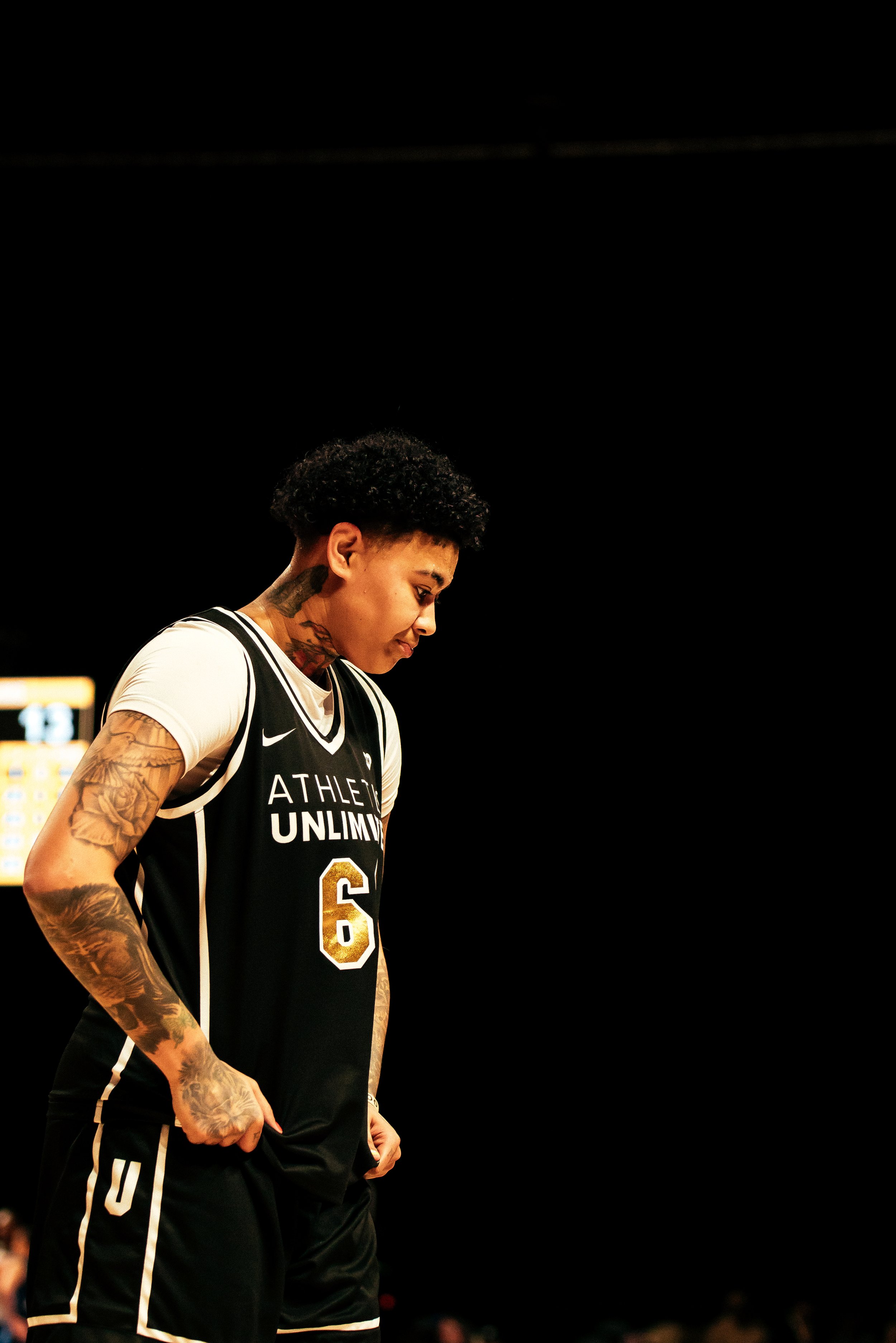 A female basketball player wearing a black jersey with the number 6 and the inscription "Athletes Unlimited" stands on a basketball court with her head bowed and hands in her pockets.