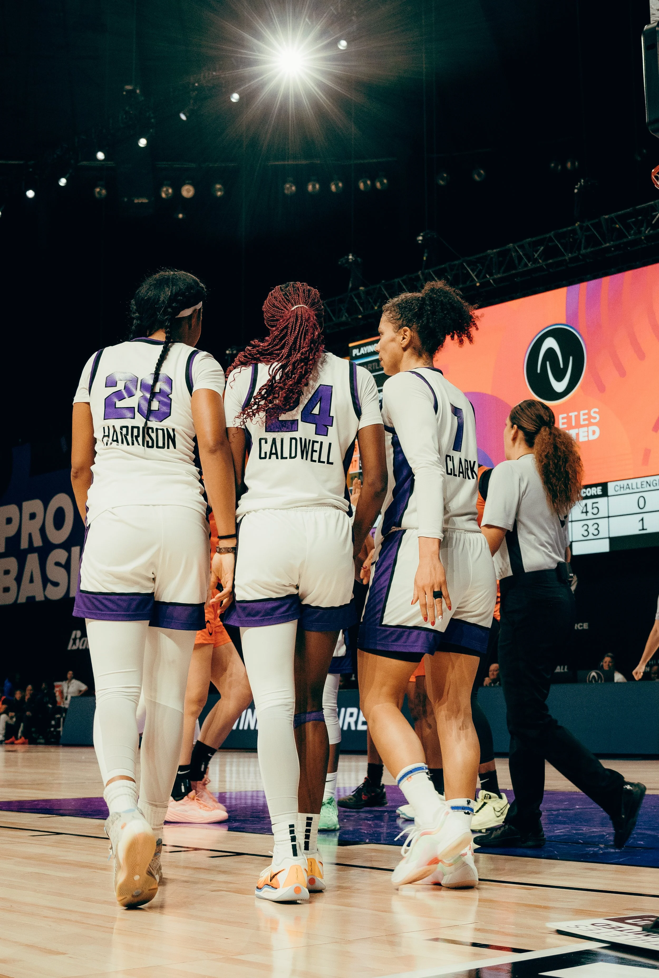 Group of female basketball players in white and purple uniforms huddling on the court during a game, with a scoreboard and bright lights overhead.