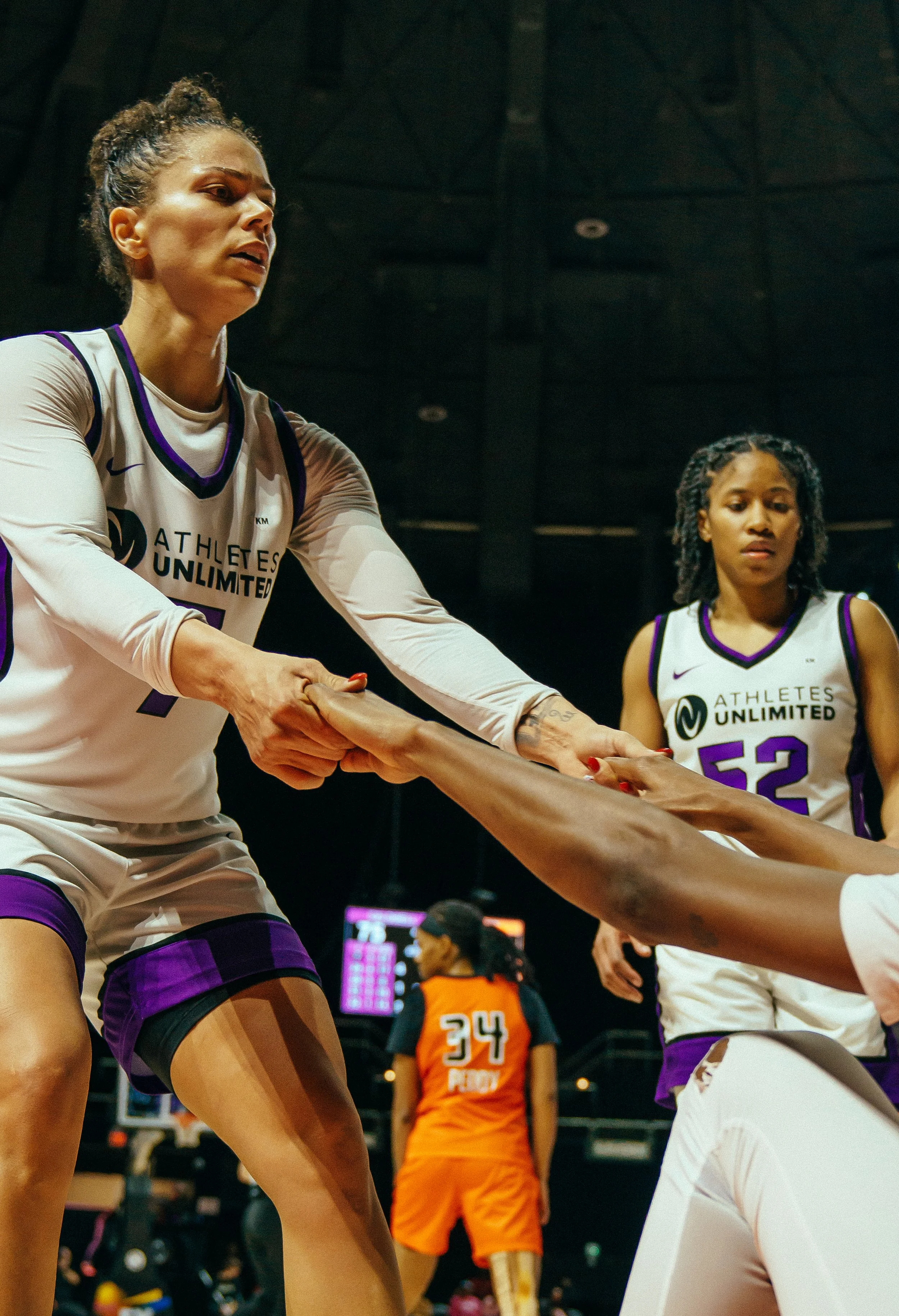 Two female basketball players from Athletes Unlimited holding hands during a game, with an opposing player in orange in the background.
