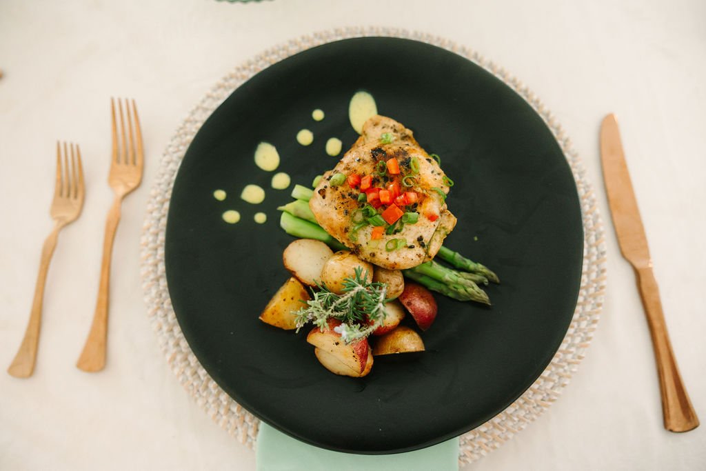 A table with two white plates of food and a vase of pink, purple, and blue flowers. One dish appears to be a piece of salmon topped with herbs and lemon, garnished with cilantro, with sauce drops around it. The other dish is a roasted chicken leg with grilled asparagus, melted cheese, and roasted tomatoes, with sauce drops around it.