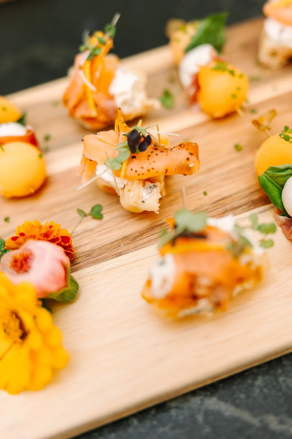 Assortment of food cups with shrimp, chicken, and slices of meat, garnished with lemon slices and tropical fruit, arranged on a wooden surface with pink flowers, and a colorful, tropical-themed card in the background.