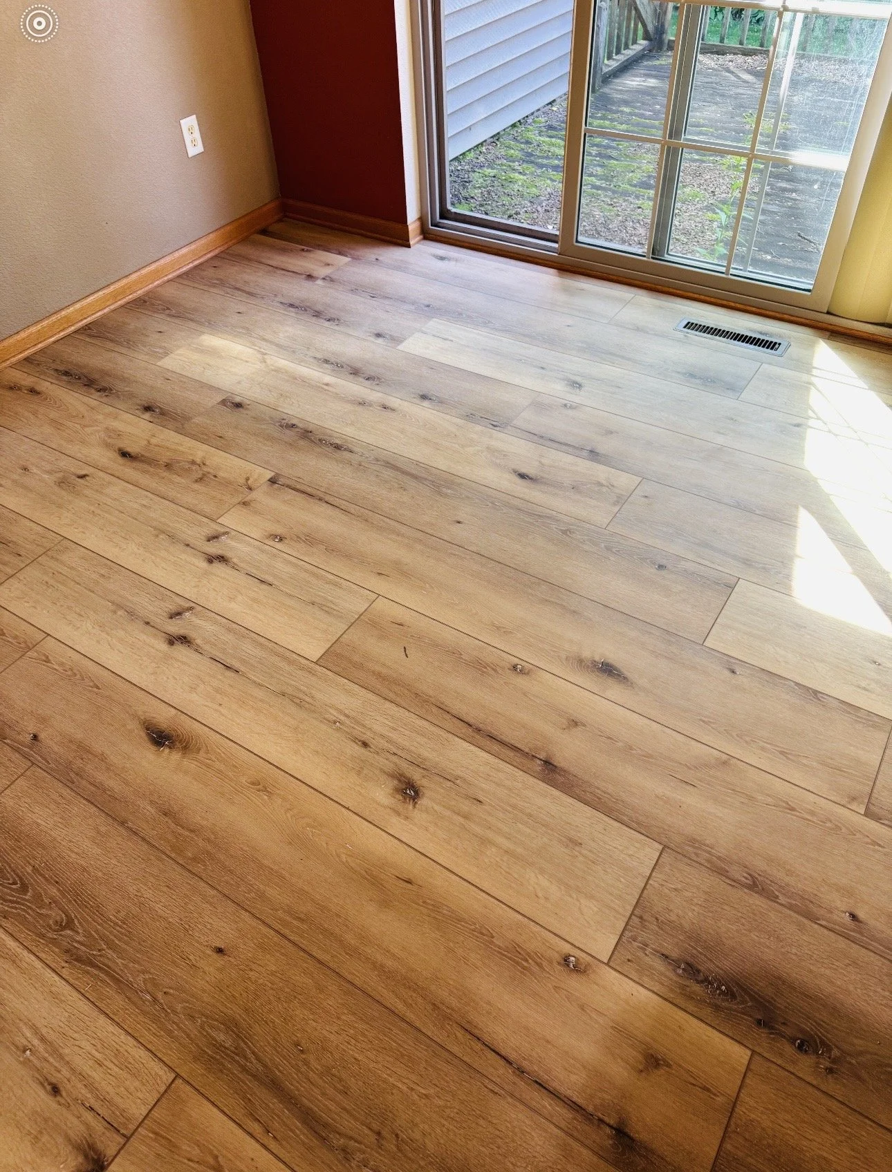 Photo of a room with light wood plank flooring, a beige wall on the left, a red accent wall in the corner, and a sliding glass door leading outside. Sunlight is shining on the floor near the door.