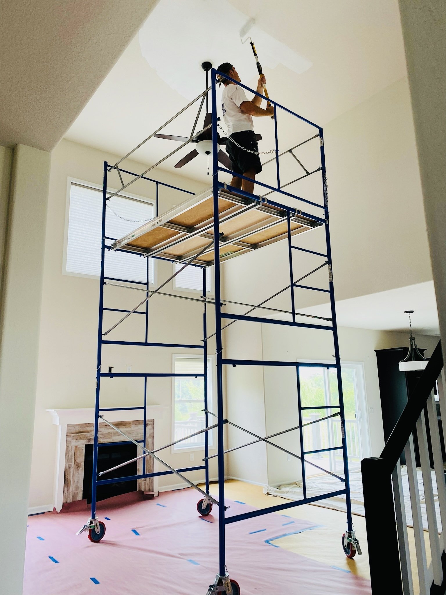 A person is standing on a blue scaffolding inside a living room, painting the ceiling with a roller.