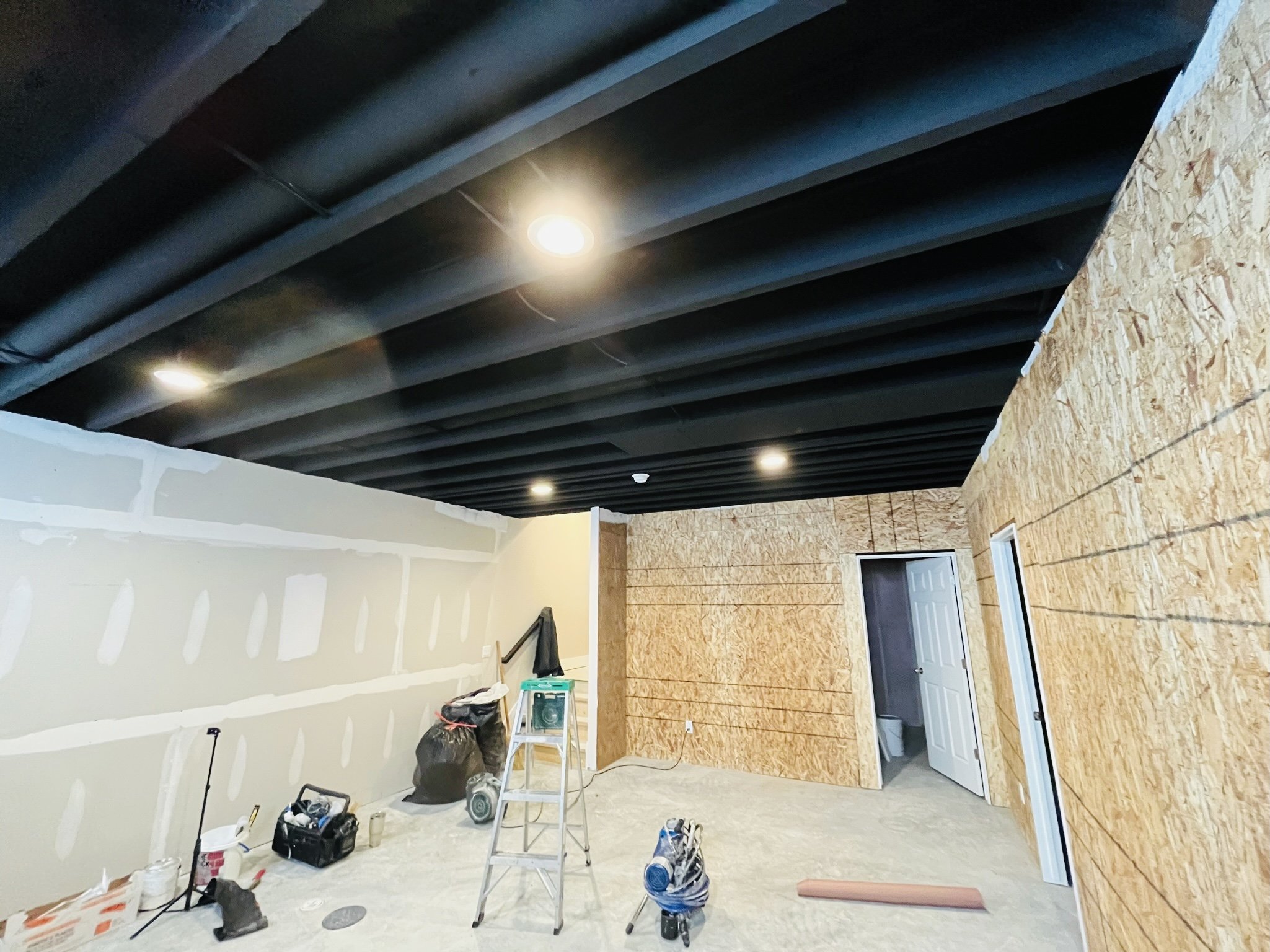Interior room under renovation with unfinished drywall and plywood walls, black ceiling with recessed lighting, construction tools and supplies on the floor.