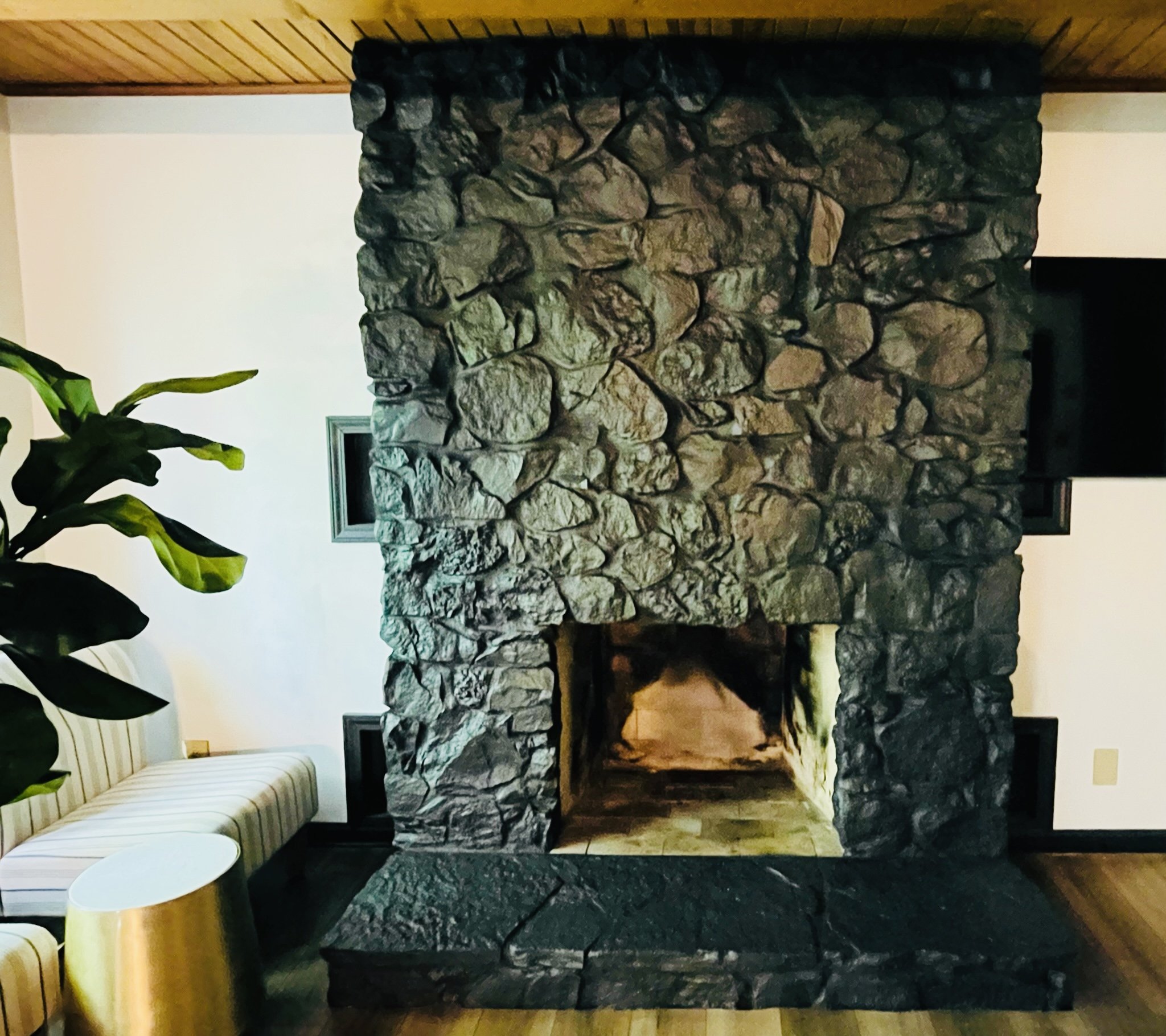 A living room features a large stone fireplace with a wood mantel, a striped armchair with a side table, a potted plant, and a wall-mounted TV.